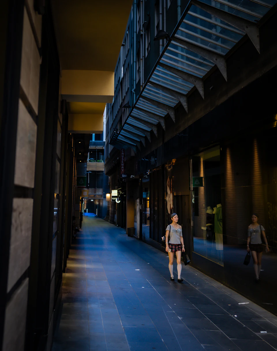 Woman walking down narrow dimly lit alleyway between tall buildings in urban setting