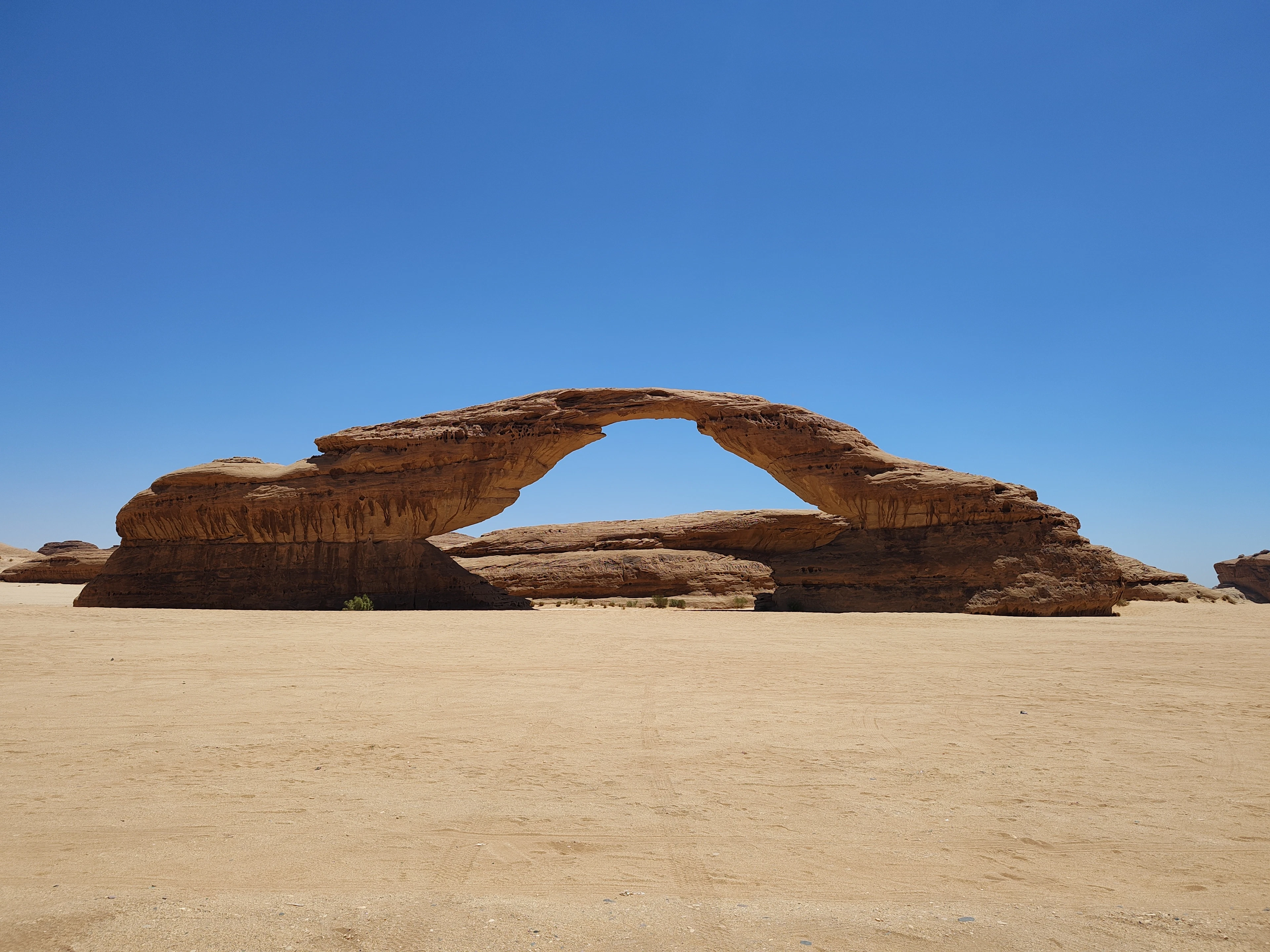 A natural sandstone rock arch stands alone on a flat desert plain under clear blue sky.
