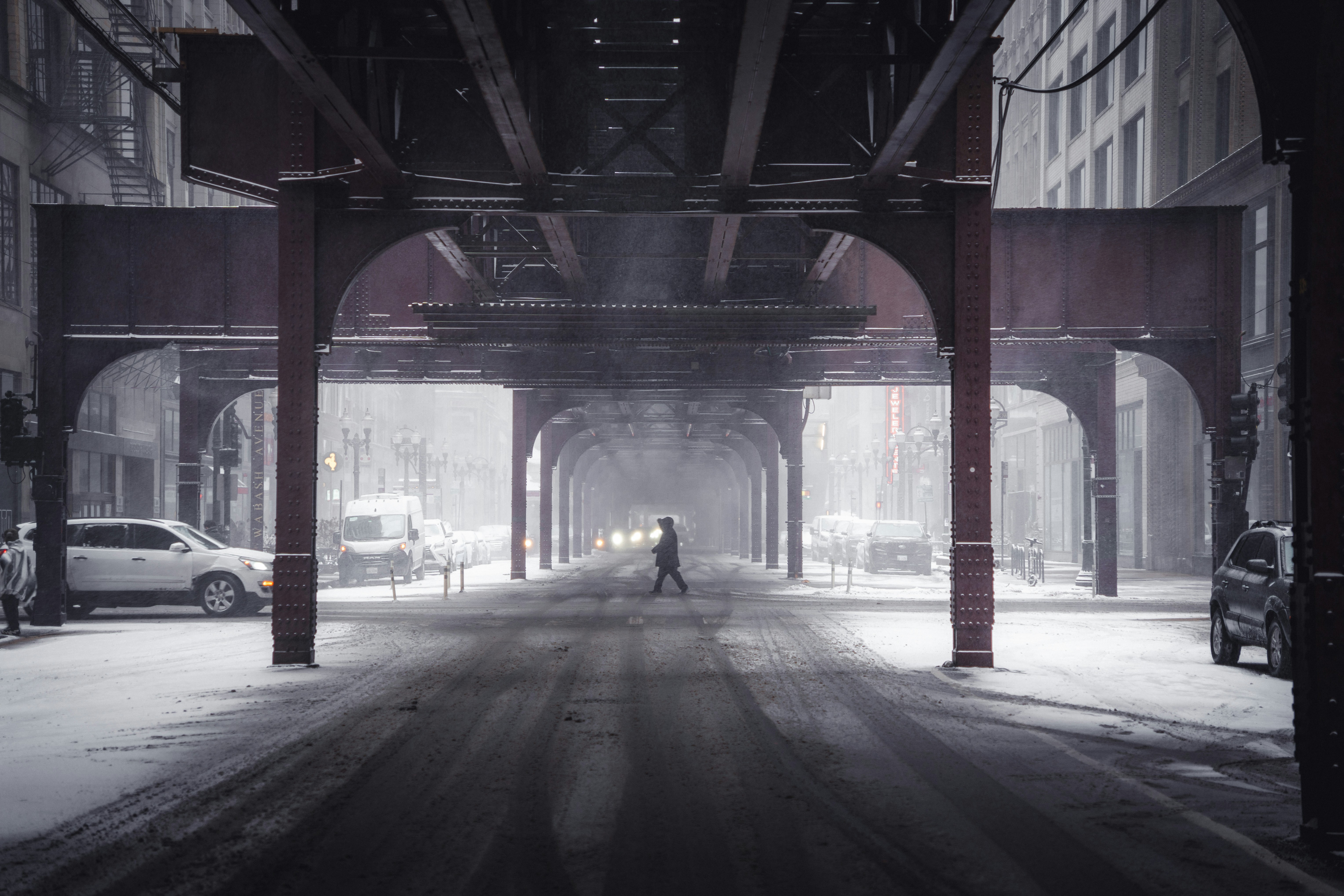 Person crossing a snowy street under elevated train tracks.