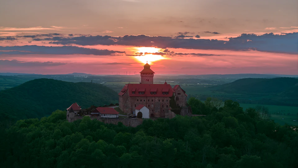 The image depicts a castle perched atop a hill, with its red roof contrasting against the sky and surrounding landscape. The sun is setting behind the castle, casting a warm glow over the scene. The castle's location on top of the hill provides an elevated vantage point, offering a panoramic view of the surrounding area.

The foreground features lush green trees that add depth to the image, while the background showcases rolling hills and fields extending into the distance. The sky above is painted in shades of orange and pink, indicating the sun's descent below the horizon.