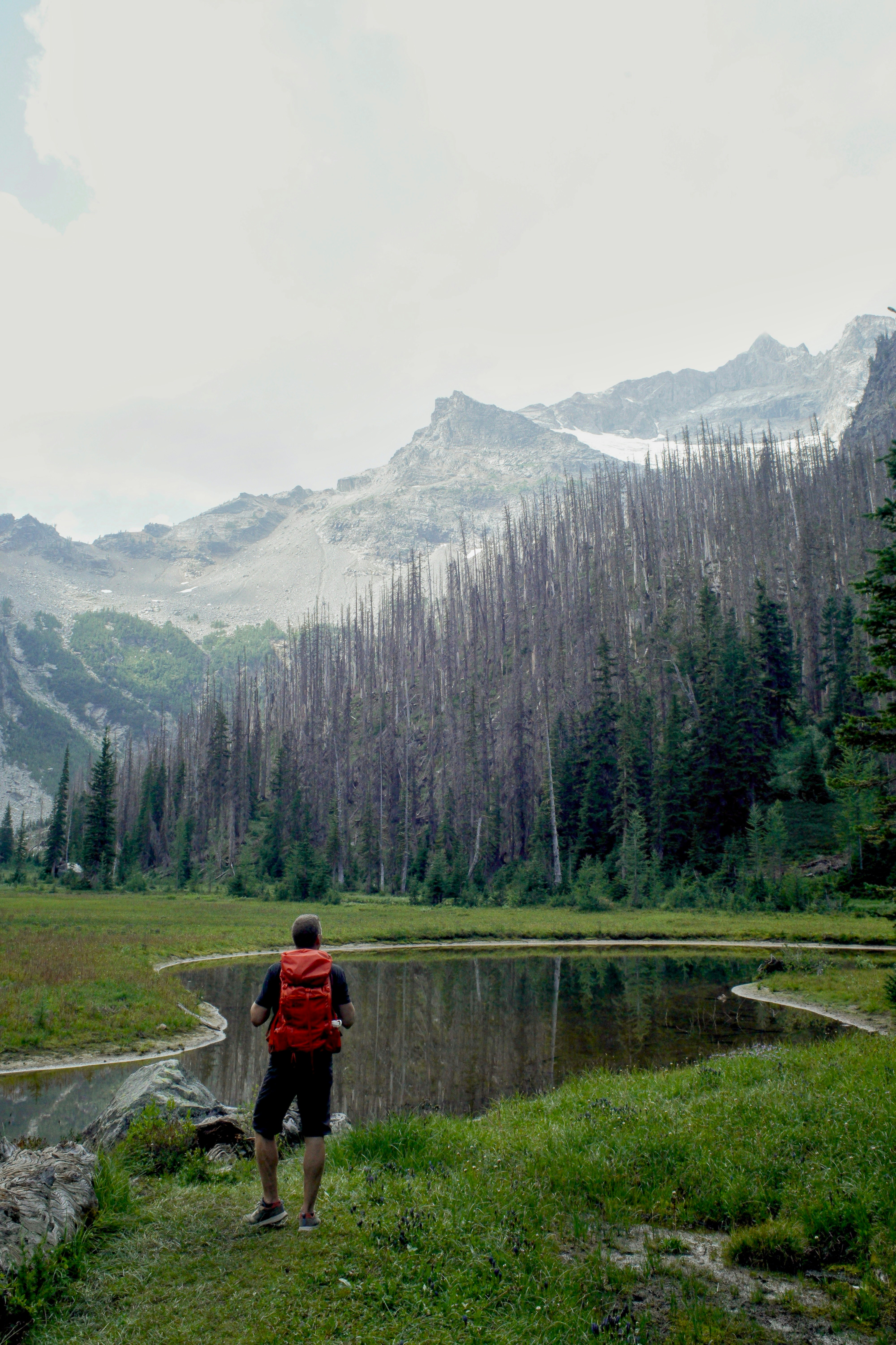 Hiker with red backpack looks at mountain lake.
