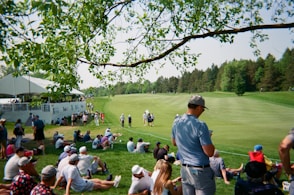 Spectators watch golfers on a sunny day at a tournament.