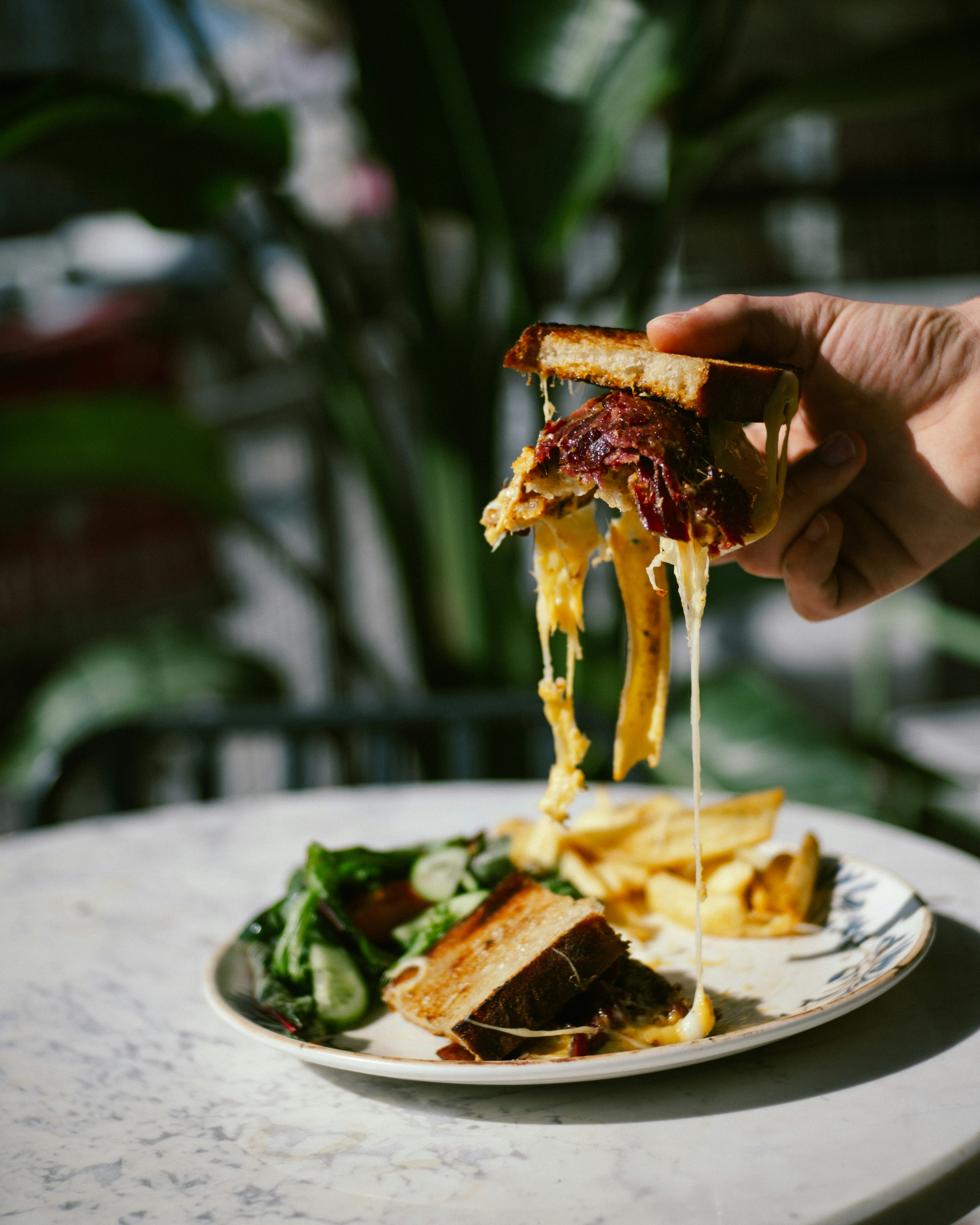Hand holding a dripping cheese sandwich over fries and salad.