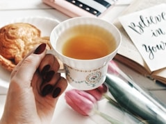Hand holding a cup of tea with pastry and flowers.