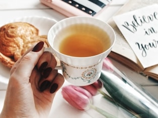Hand holding a cup of tea with pastry and flowers.