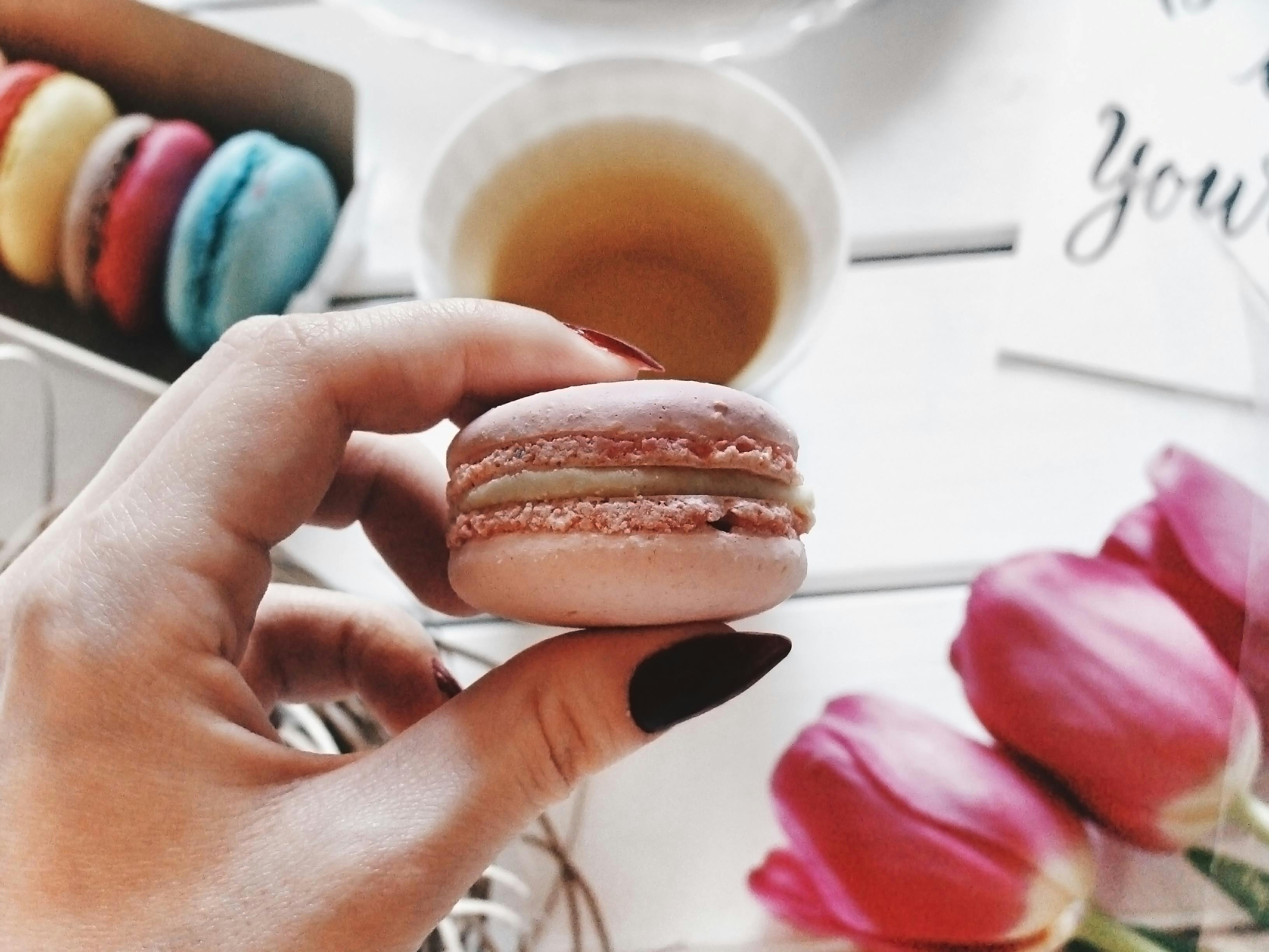 Hand holding a pink macaron with colorful macarons and tea.