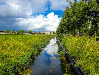 A narrow canal flows through a green landscape under clouds.