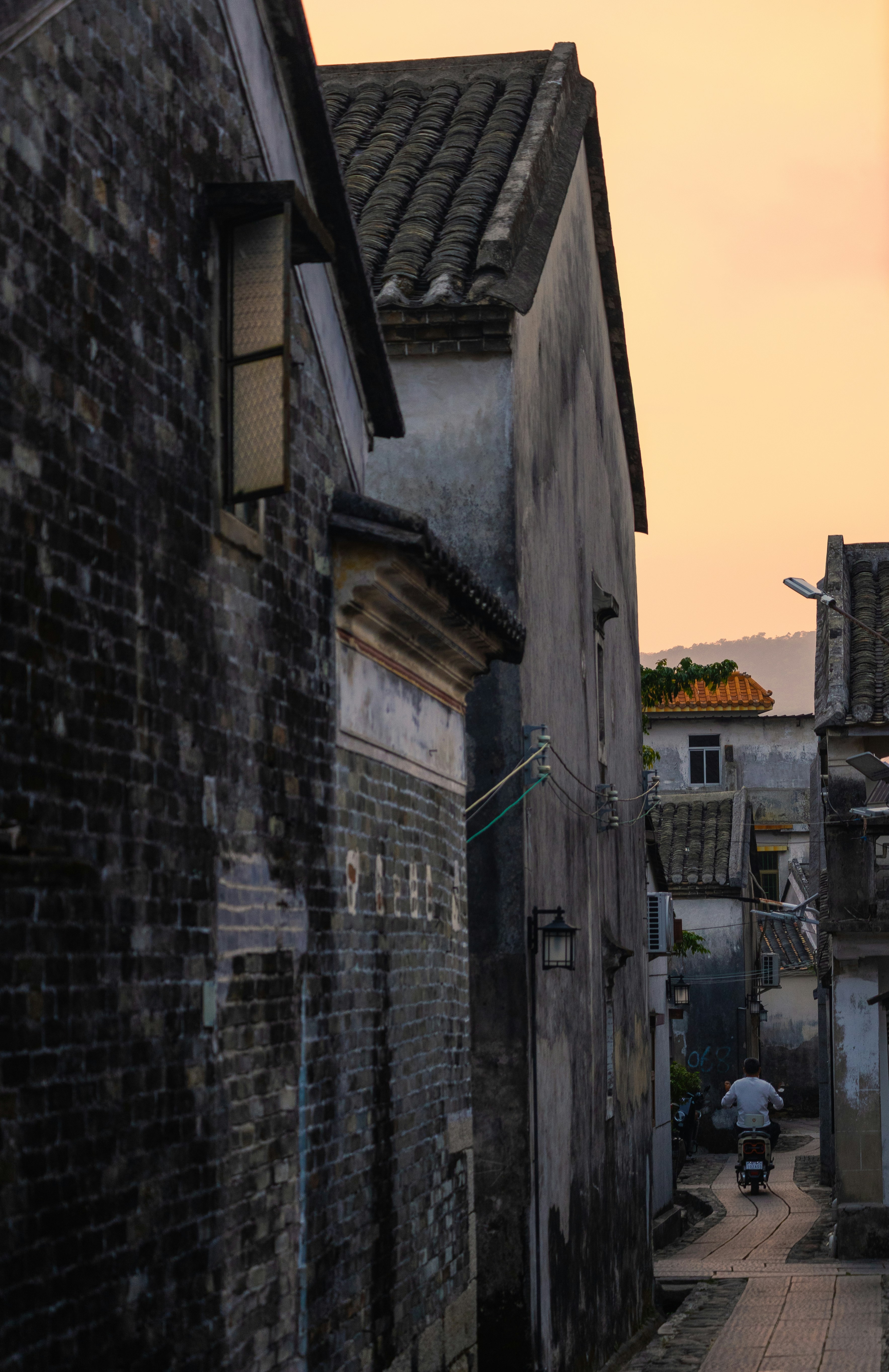 Man rides motorcycle down narrow street at dusk.