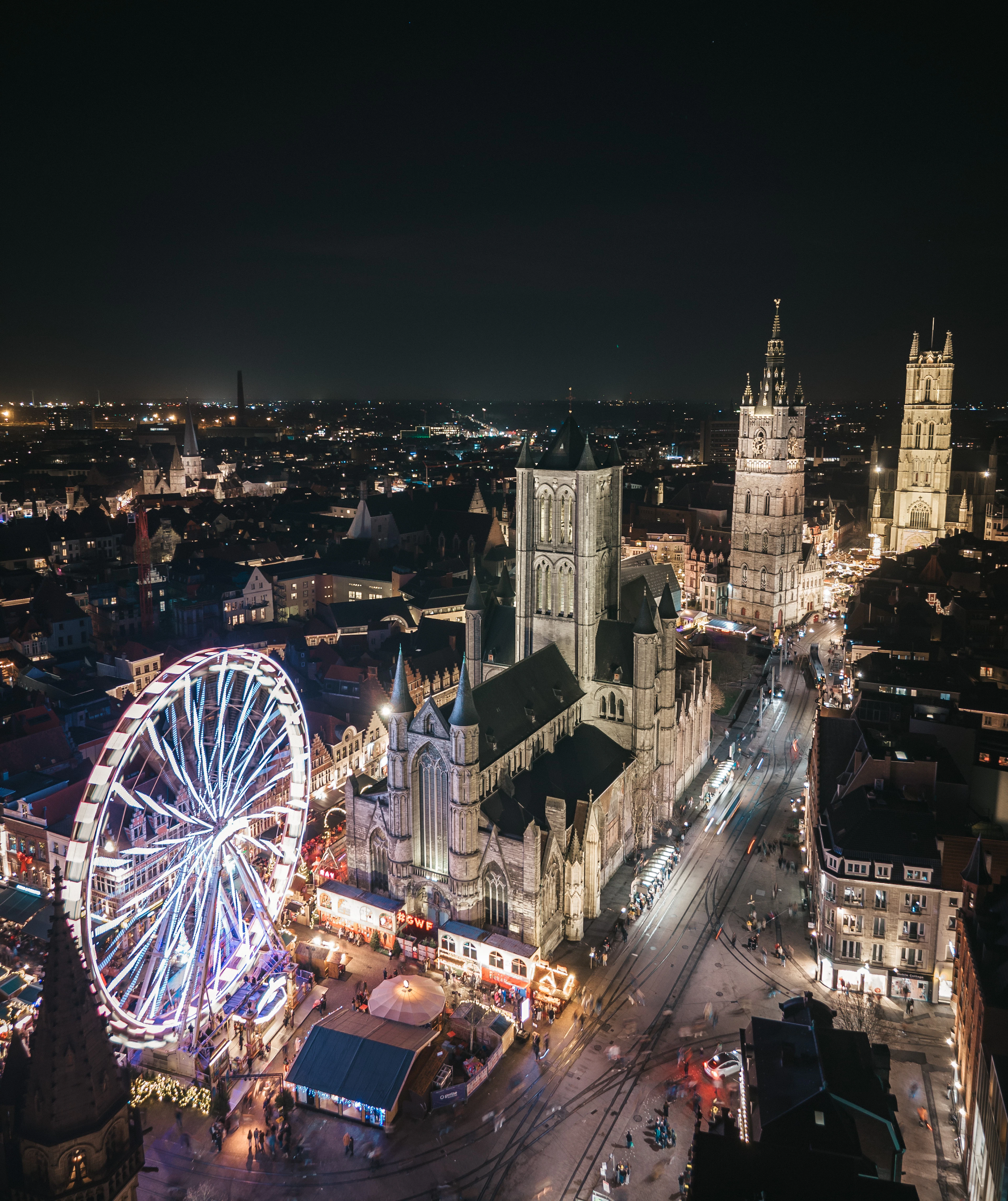 A night view over Ghent's floodlit medieval centre, a brightly lit ferris wheel and winter market stalls beside Saint Nicholas' Church, with the Belfry and the tower of Saint Bavo's Cathedral rising beyond.