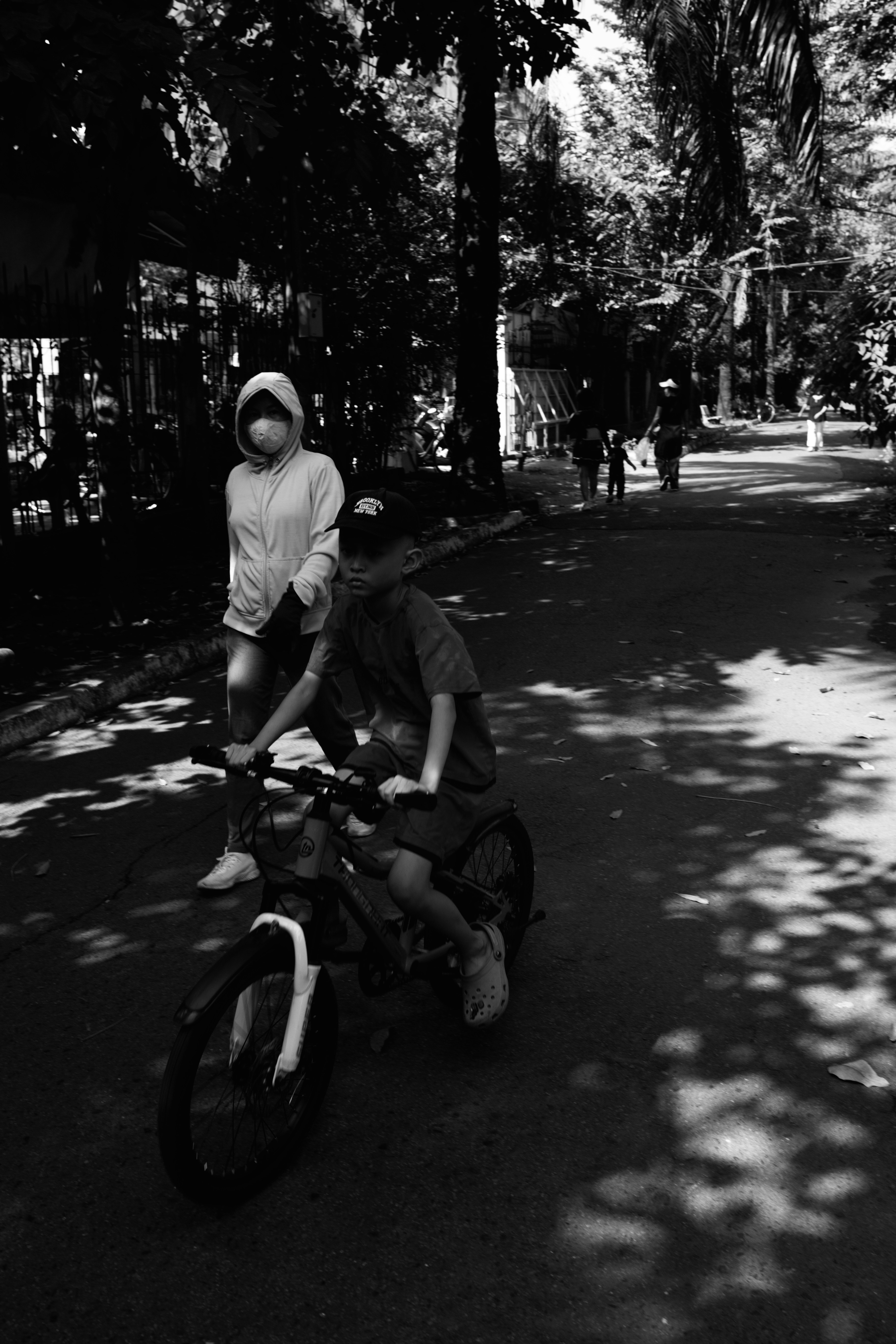 A child riding a bicycle on a tree-lined path with an adult walking behind