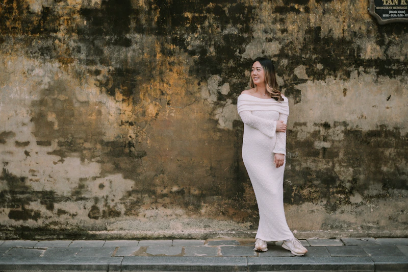 Woman in a long dress stands against a textured wall.