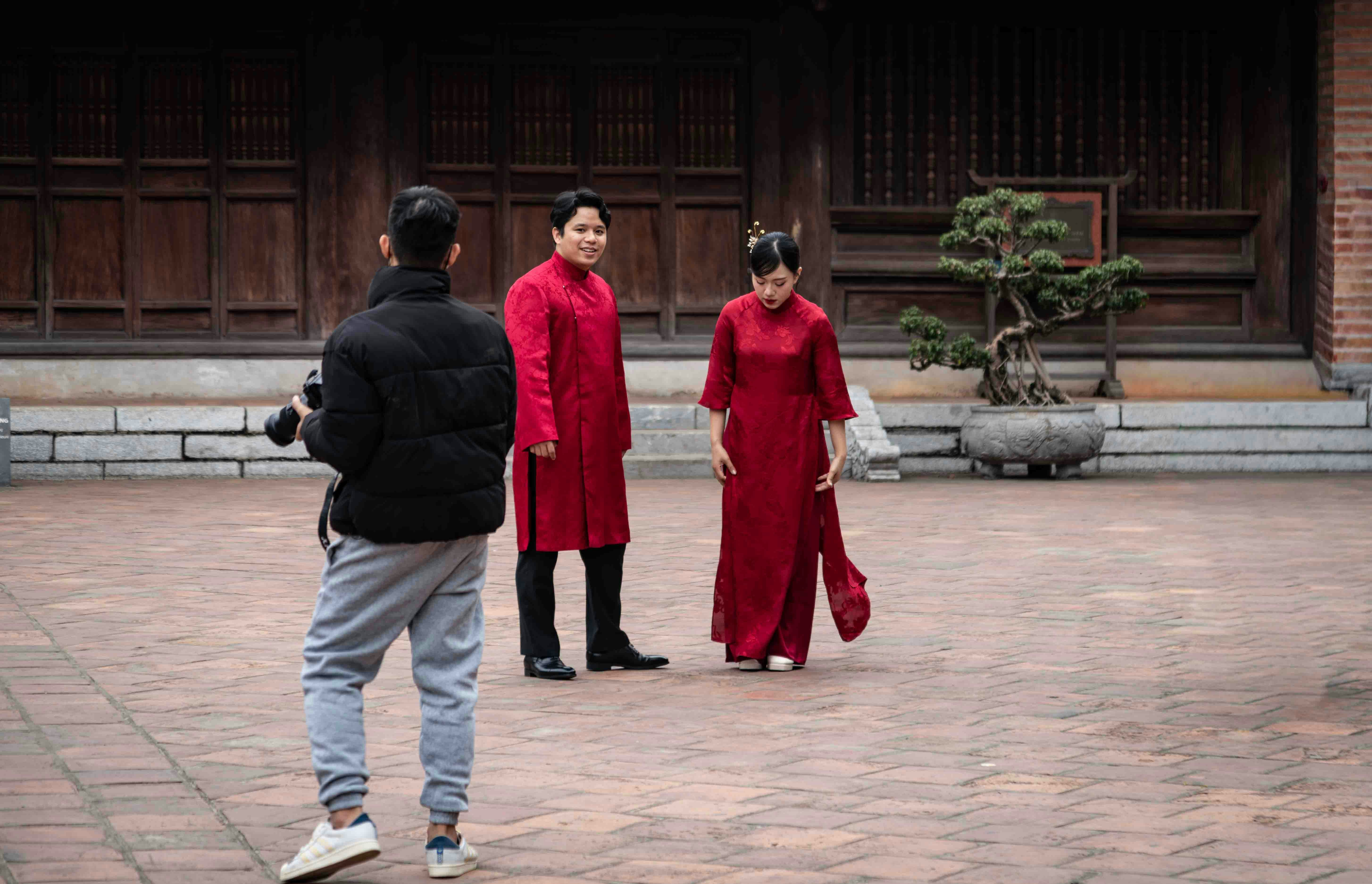 A photographer takes a picture of a man and a woman wearing Asian traditional red outfits in a courtyard with a bonsai tree