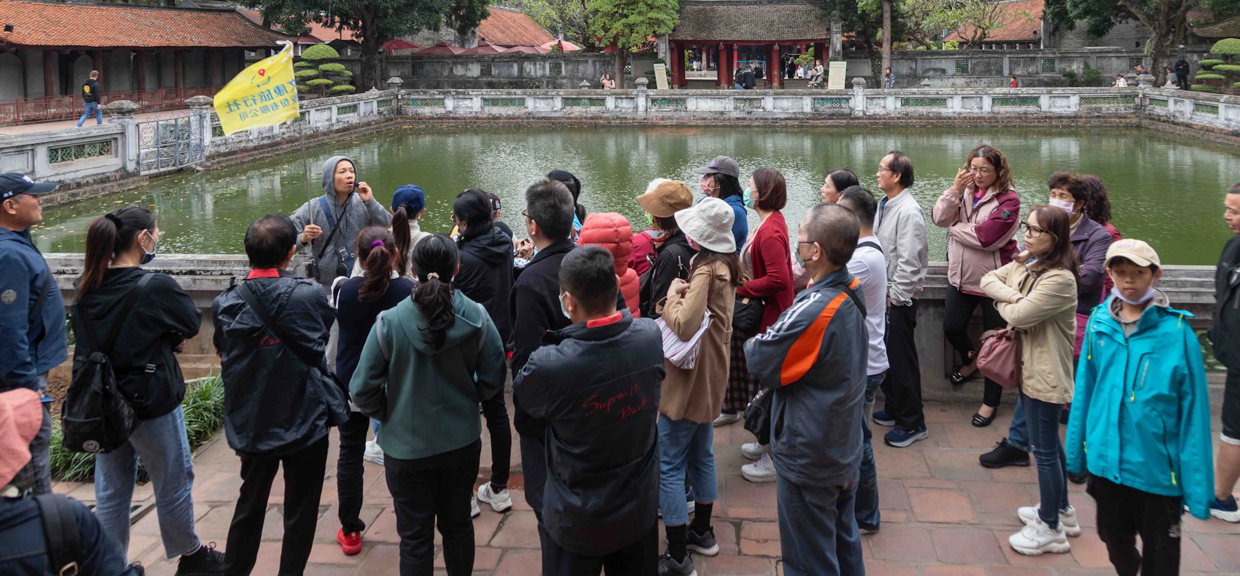 A guide holding a yellow flag speaks to a tour group beside a pond and historic buildings.
