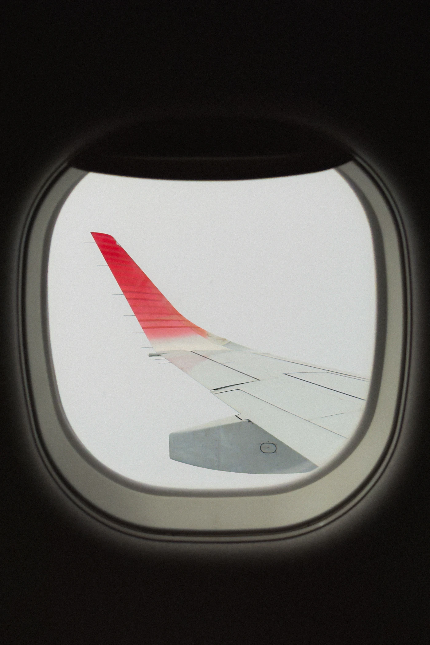 The view through a passenger window of an airplane wing with a red tip against a bright sky in flight