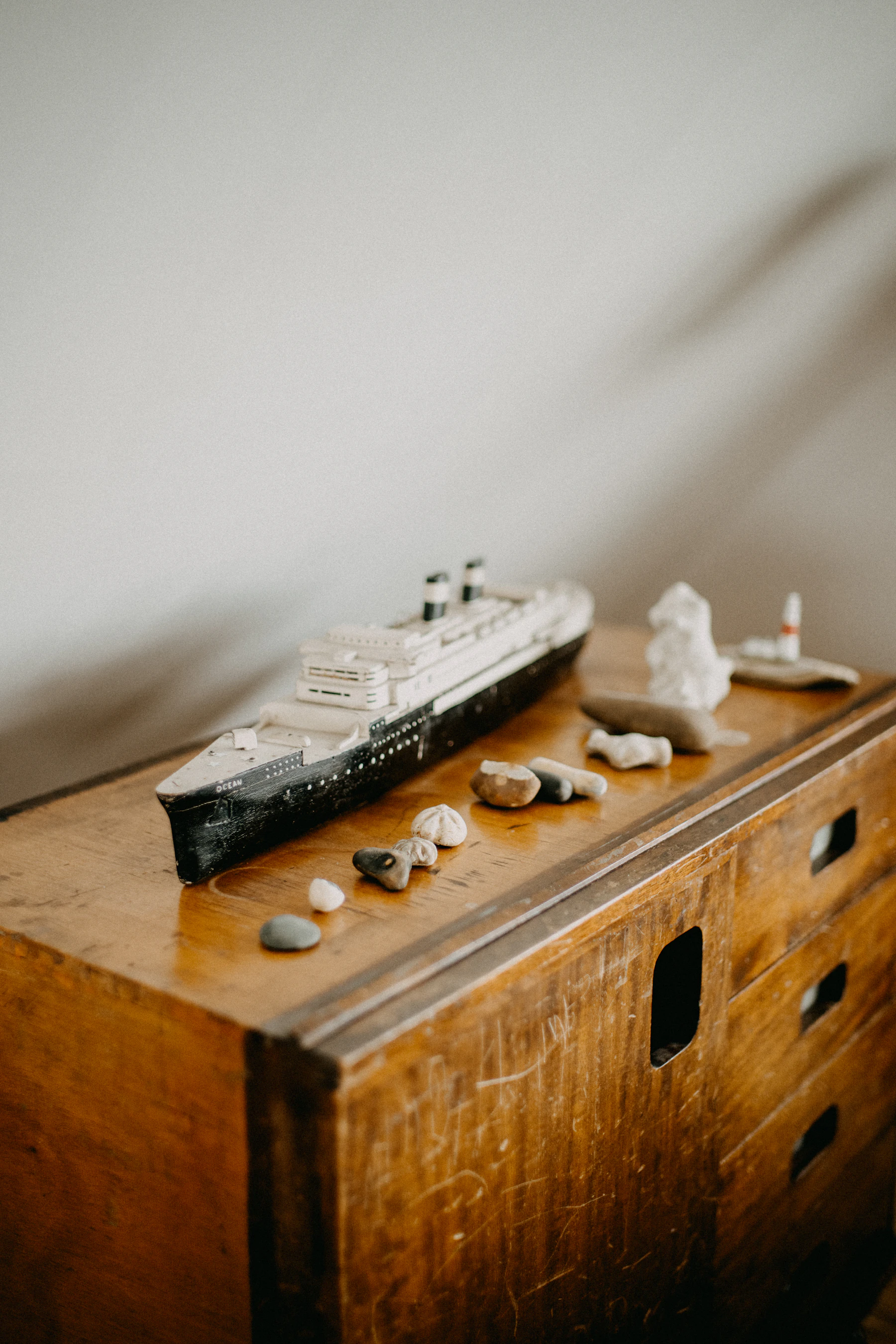 A small black and white model passenger ship on a wooden dresser beside stones and shells