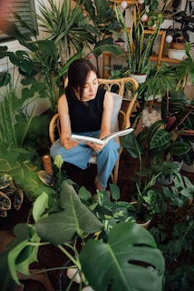 Woman reading a book surrounded by lush green plants.