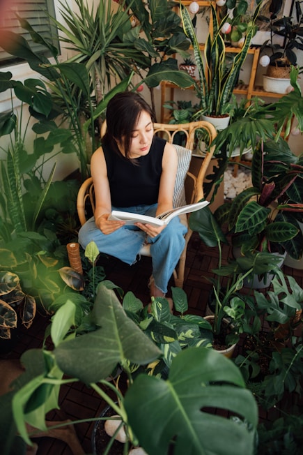 Woman reading a book surrounded by lush green plants.