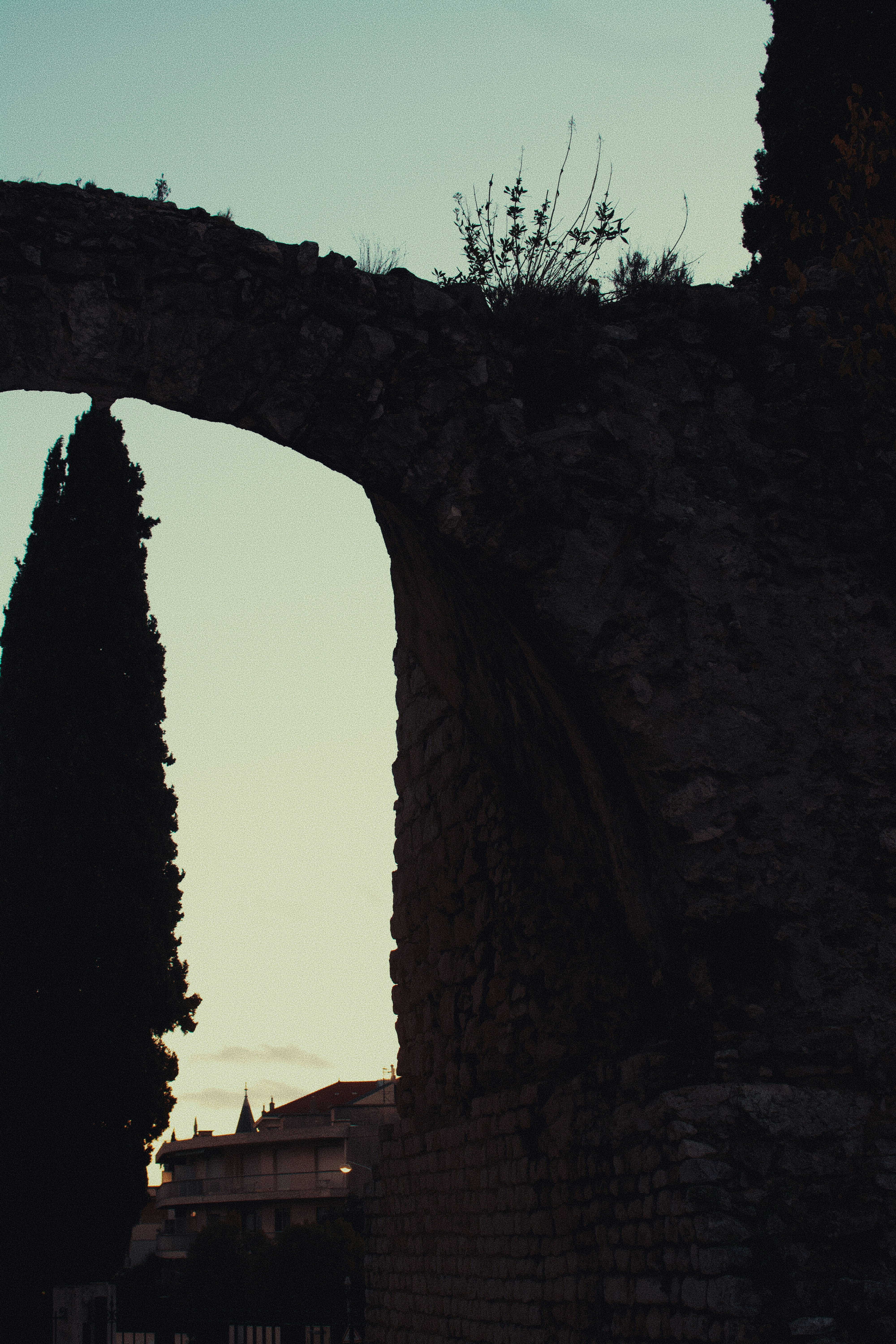 Ancient stone archway with cypress trees and buildings.