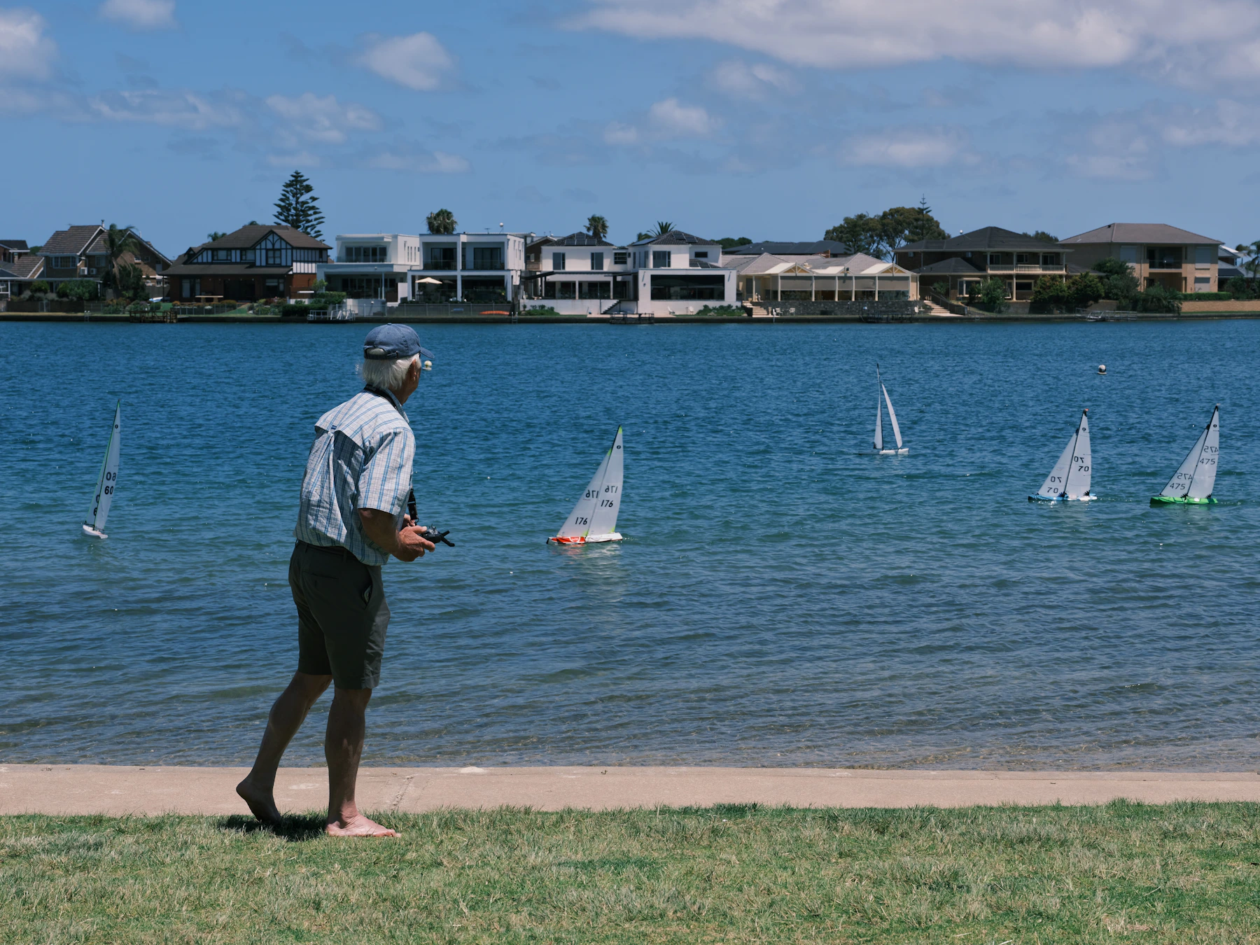 A man on a grassy bank holding a transmitter while five model sailboats race across a blue lake