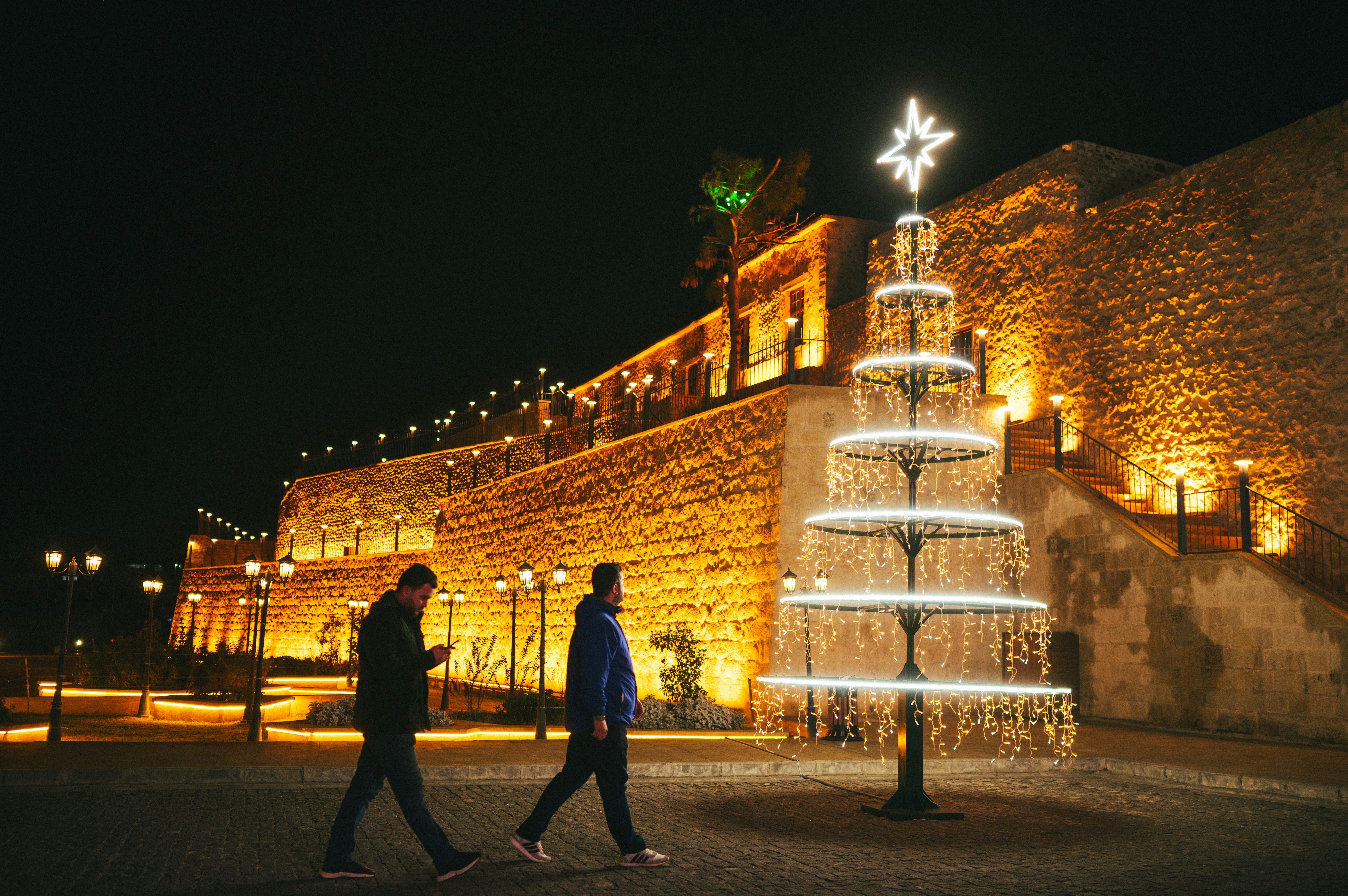Two people walk past a lit christmas tree at night.