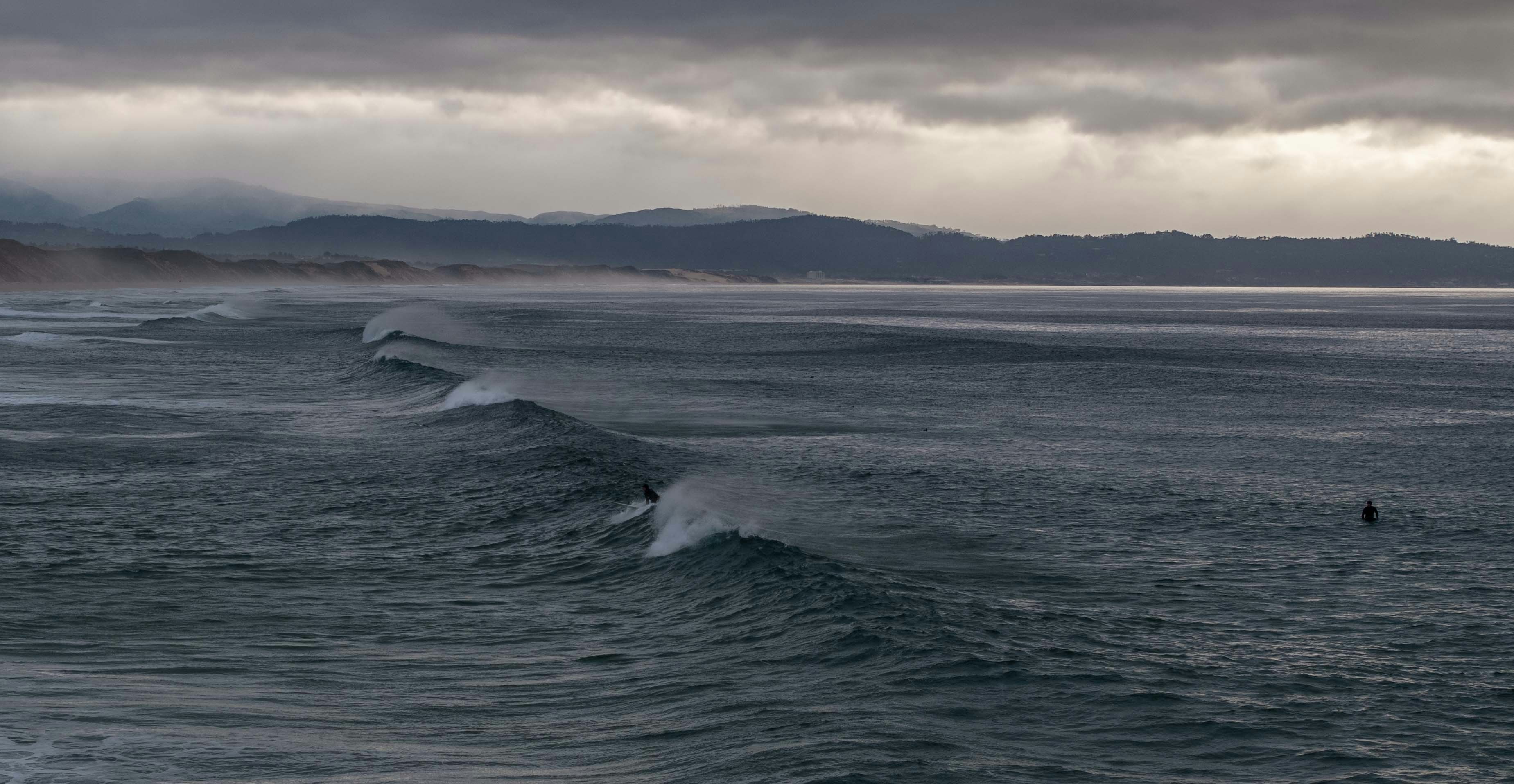 Waves roll onto a misty, overcast beach with a surfer.