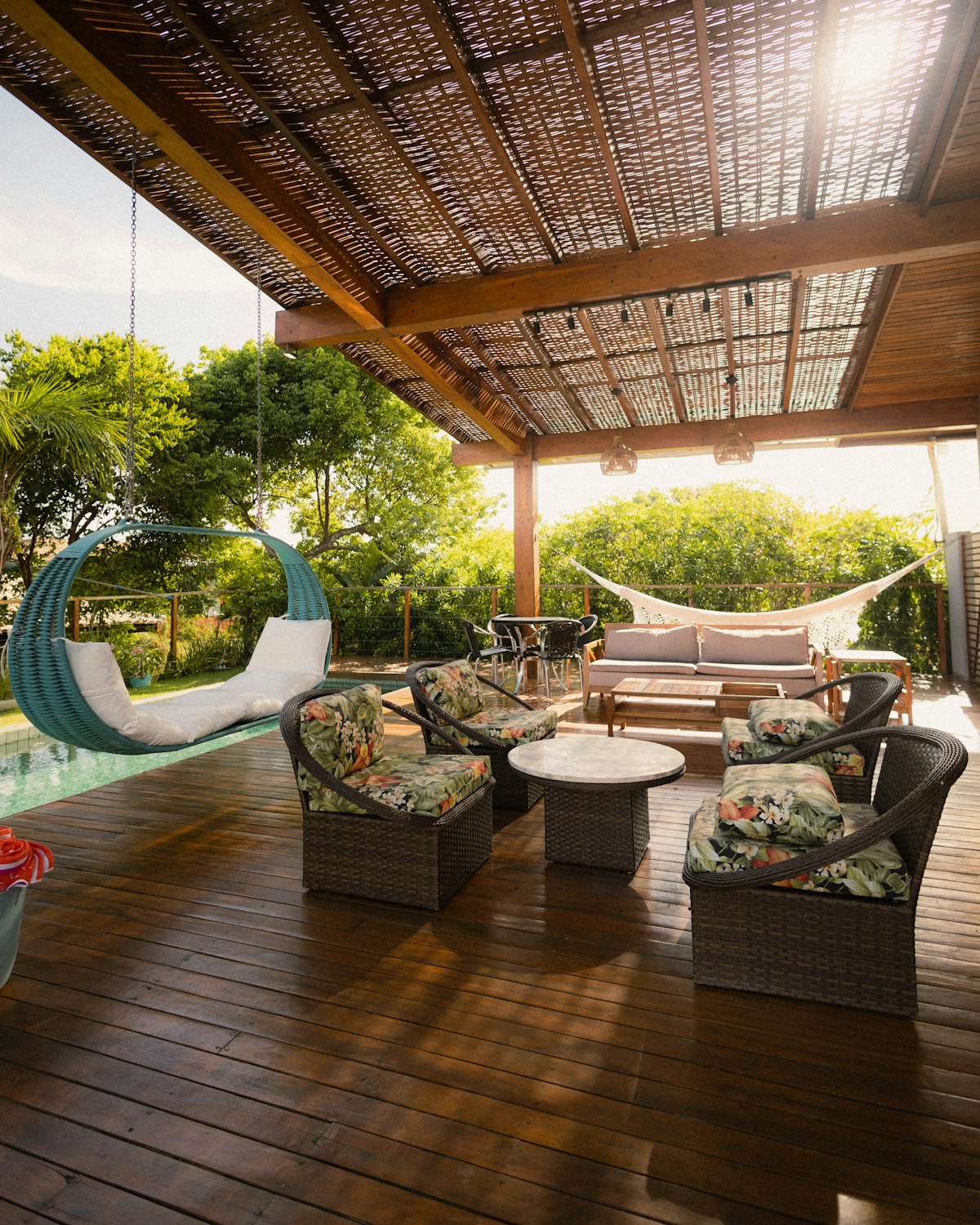 Pérgola de madera con techo de cañizo sobre un deck, con sillones de ratán con almohadones floreados, un sillón colgante turquesa y una hamaca paraguaya al fondo