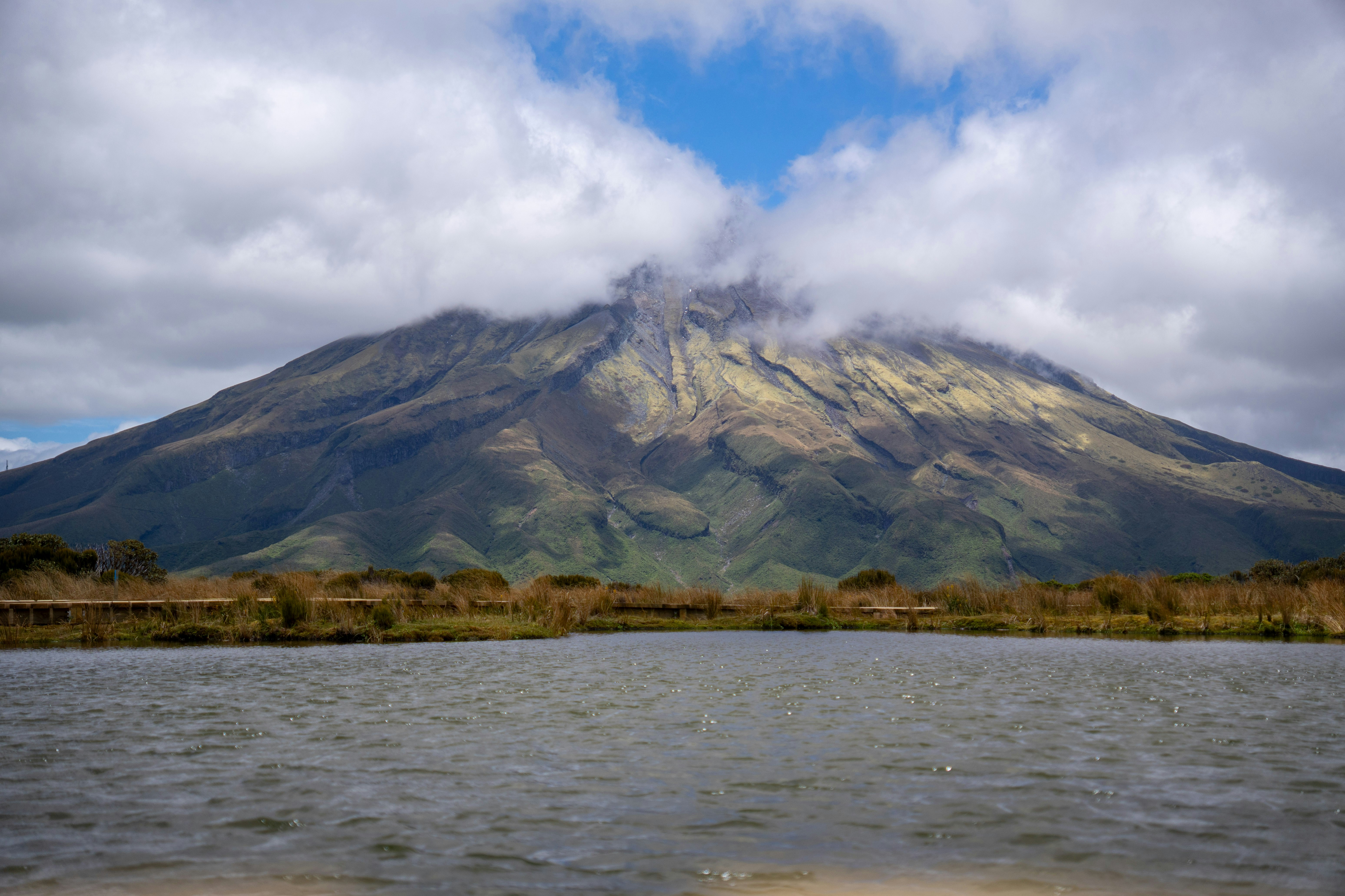 Volcano shrouded in clouds with a lake in foreground.