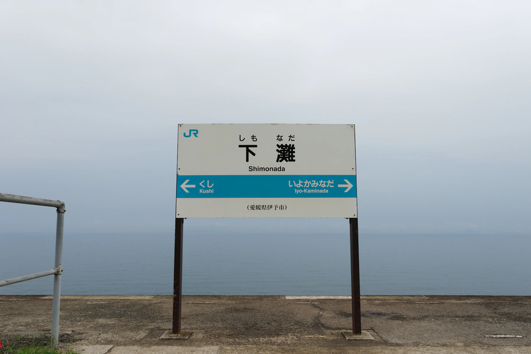 A white station name board standing on an open platform beside a grey, misty sea
