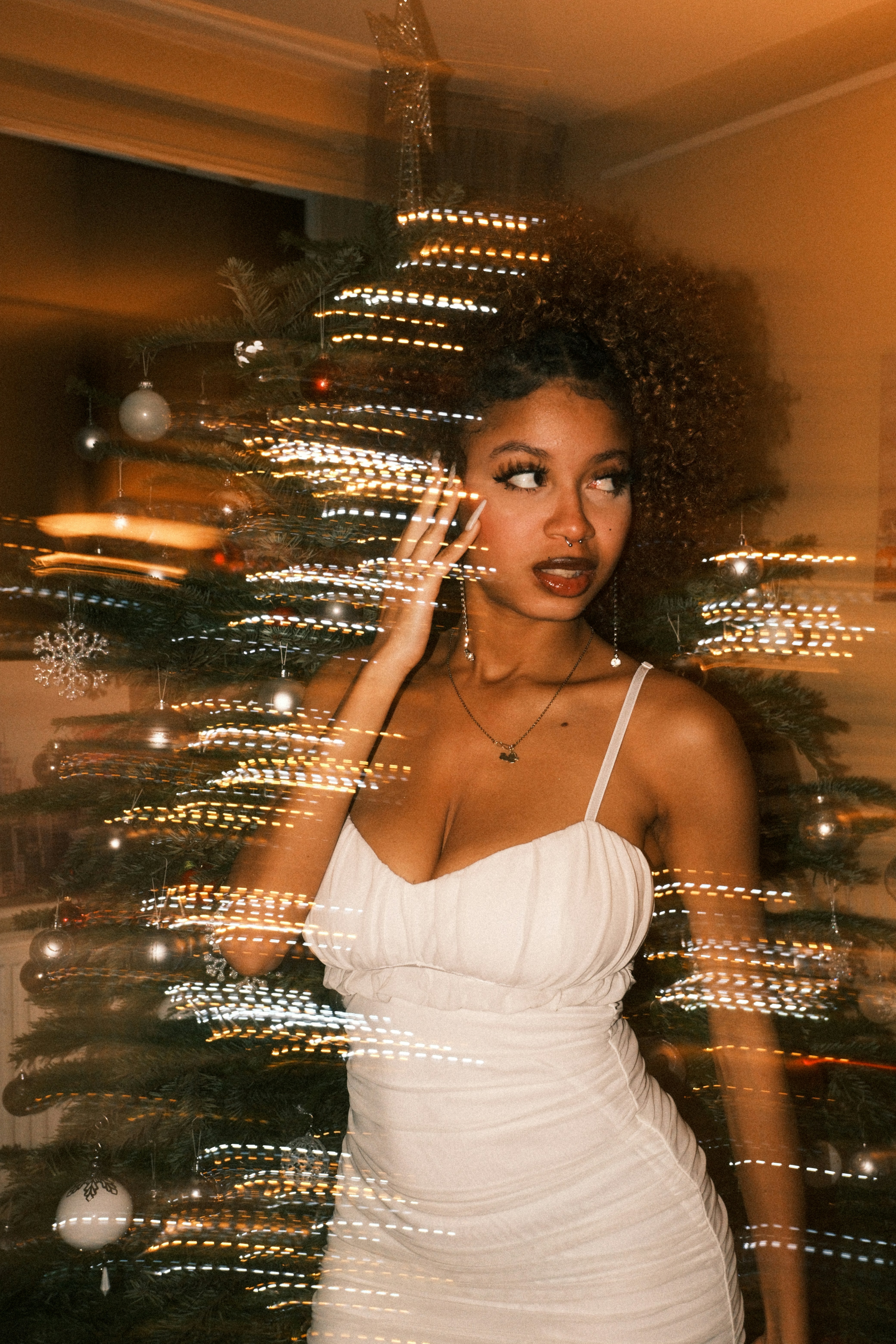 Young woman in white dress near blurred christmas tree lights.