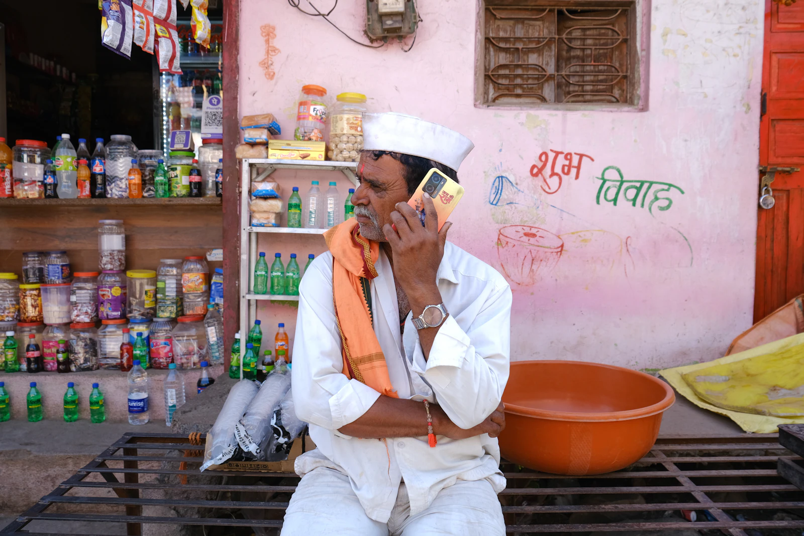 A shopkeeper on a phone call inside a small corner shop, bottles stacked behind him