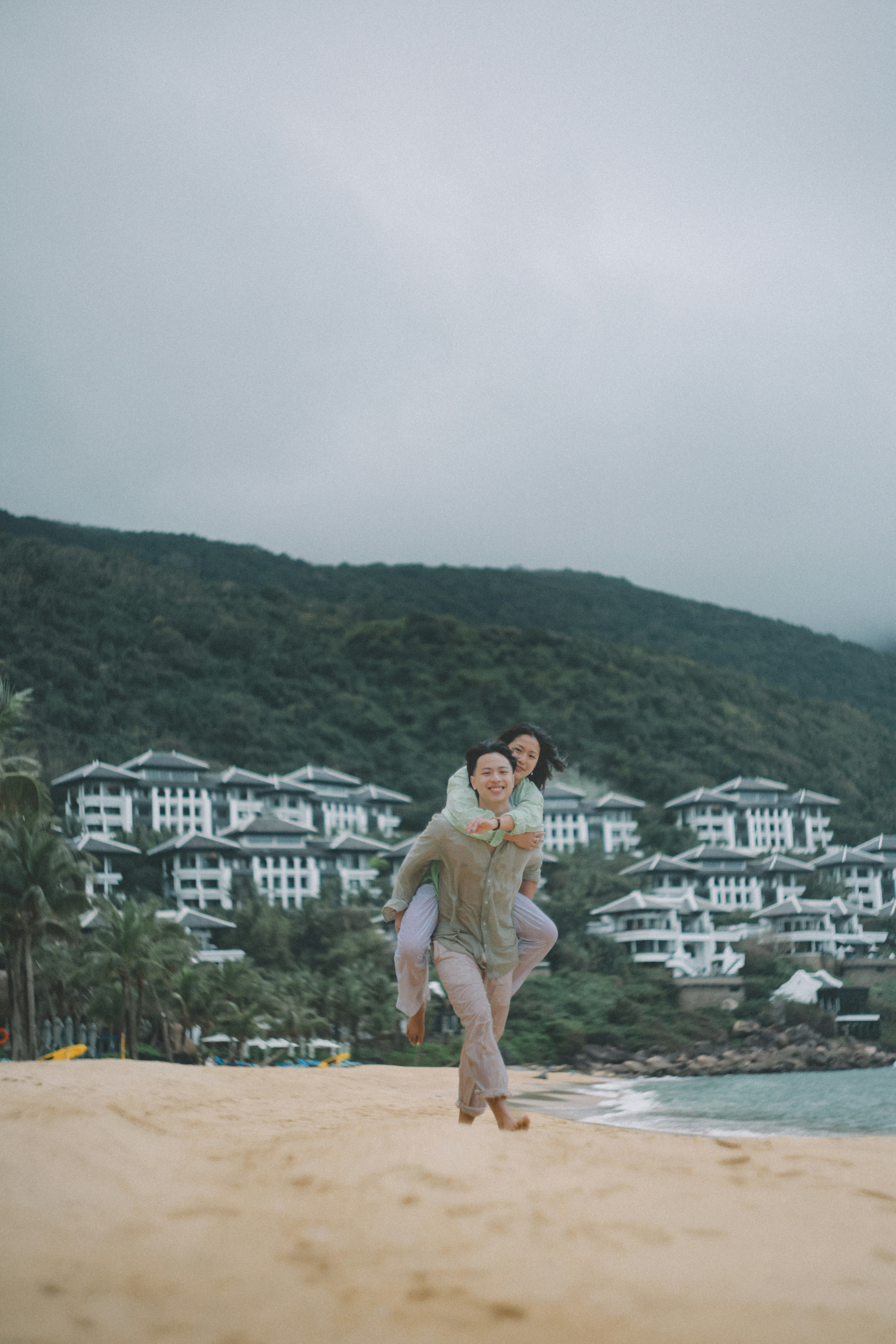 Couple piggybacking on a sandy beach with resort buildings.