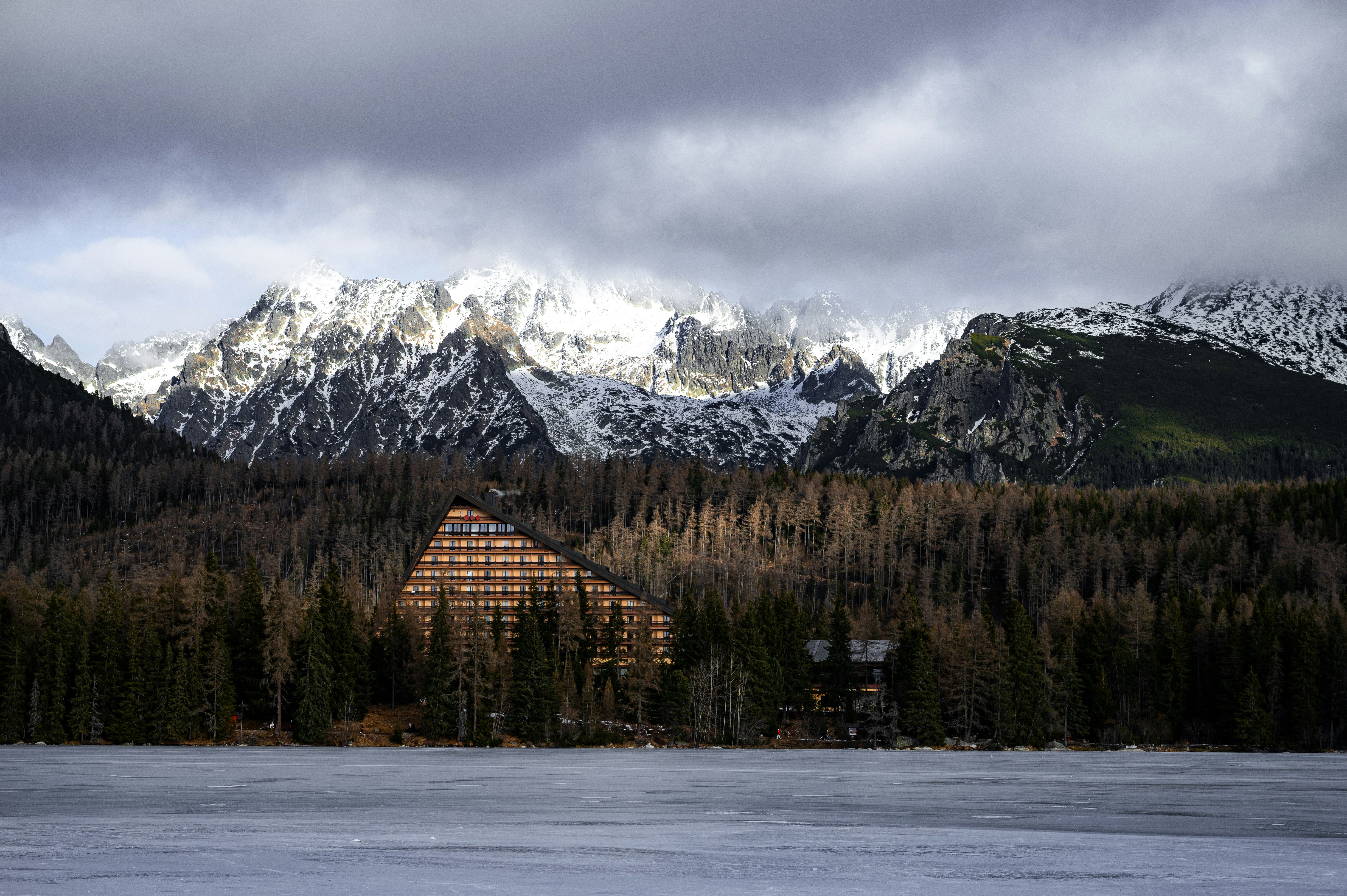 Wooden lodge nestled among trees before snowy mountains.