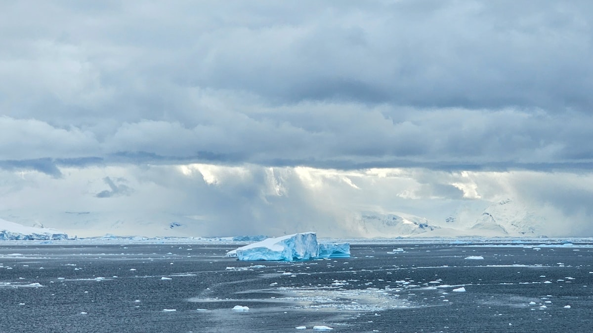 一座平顶的蓝白色冰山浮在散布着碎冰的深灰色海面上，地平线上是覆雪的山脉，几道光束穿透厚重的云层