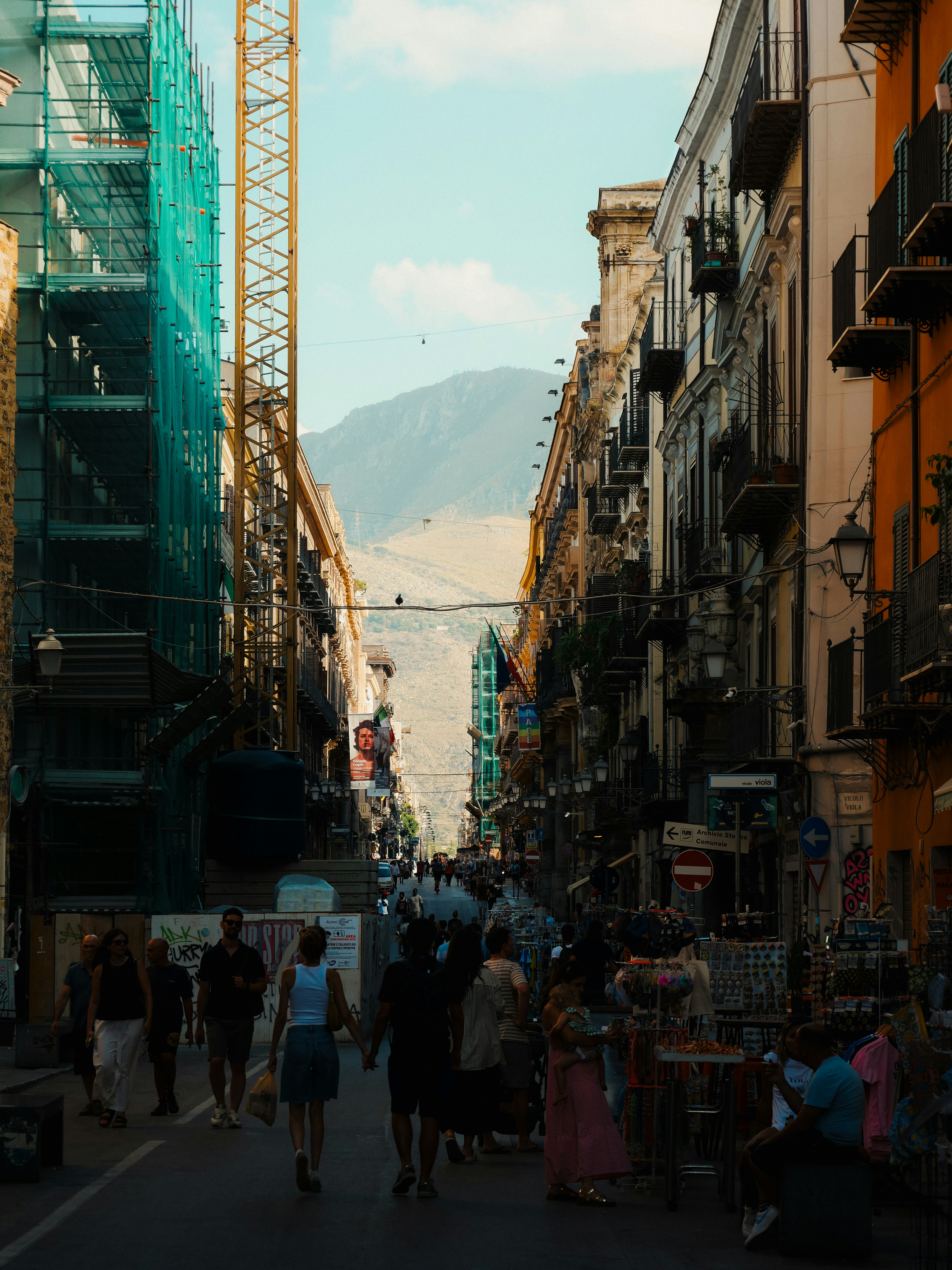 People walk down a narrow street with buildings and mountain.