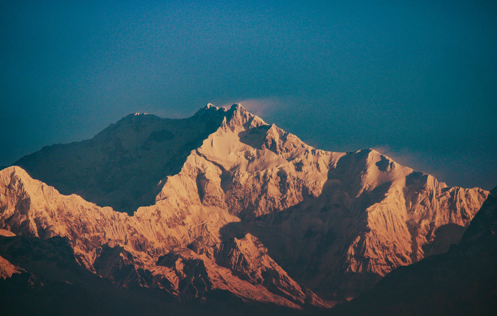 The summit ridges of Kanchenjunga lit gold by the rising sun against a deep blue dawn sky, the lower flanks still in shadow