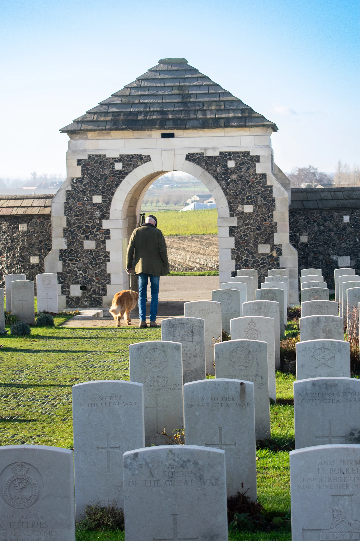 A man walks slowly with his dog through the gate of a quiet war cemetery in soft daylight, the two of them side by side and unhurried