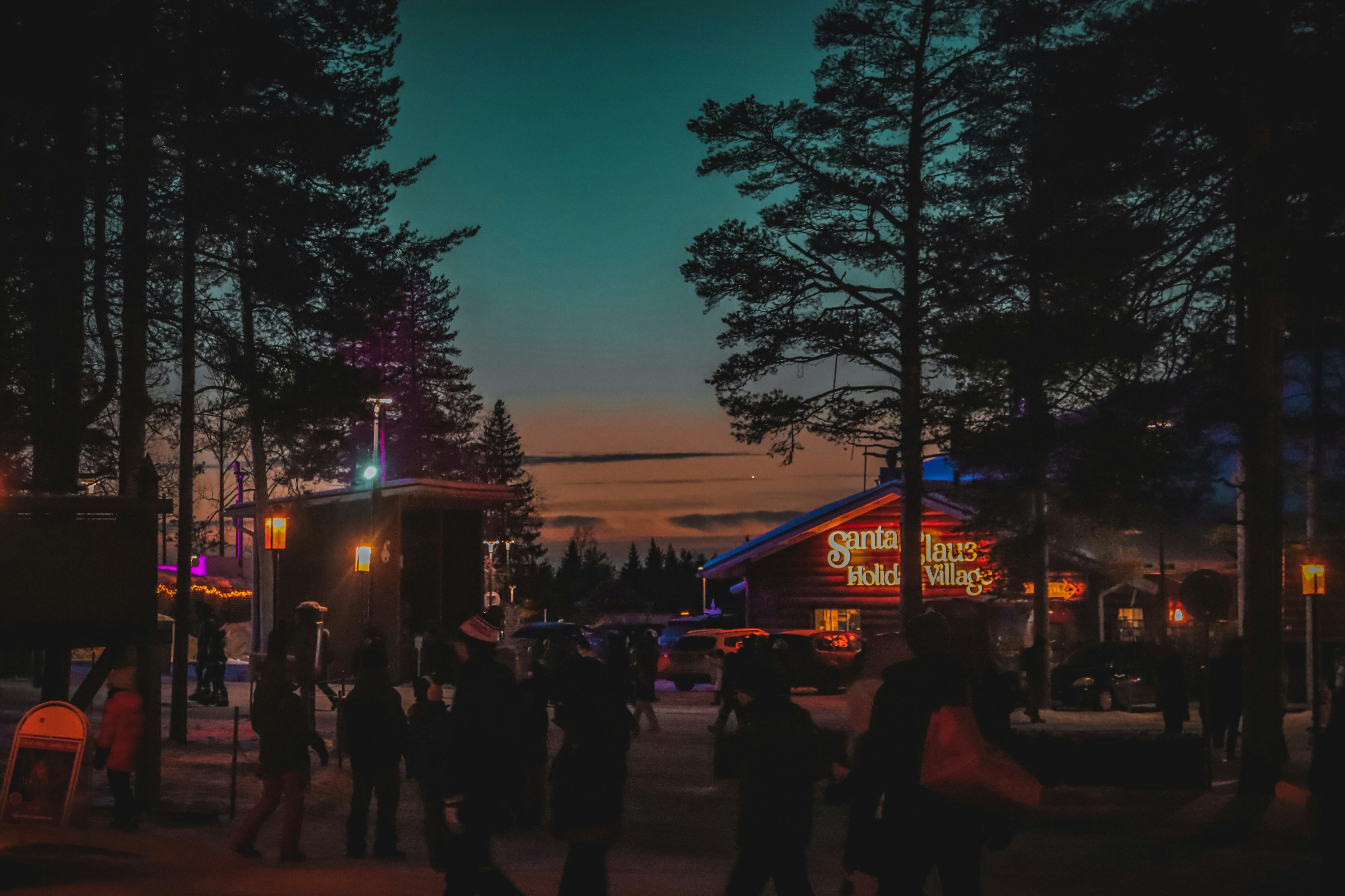 People walk through a village at dusk with colorful lights.