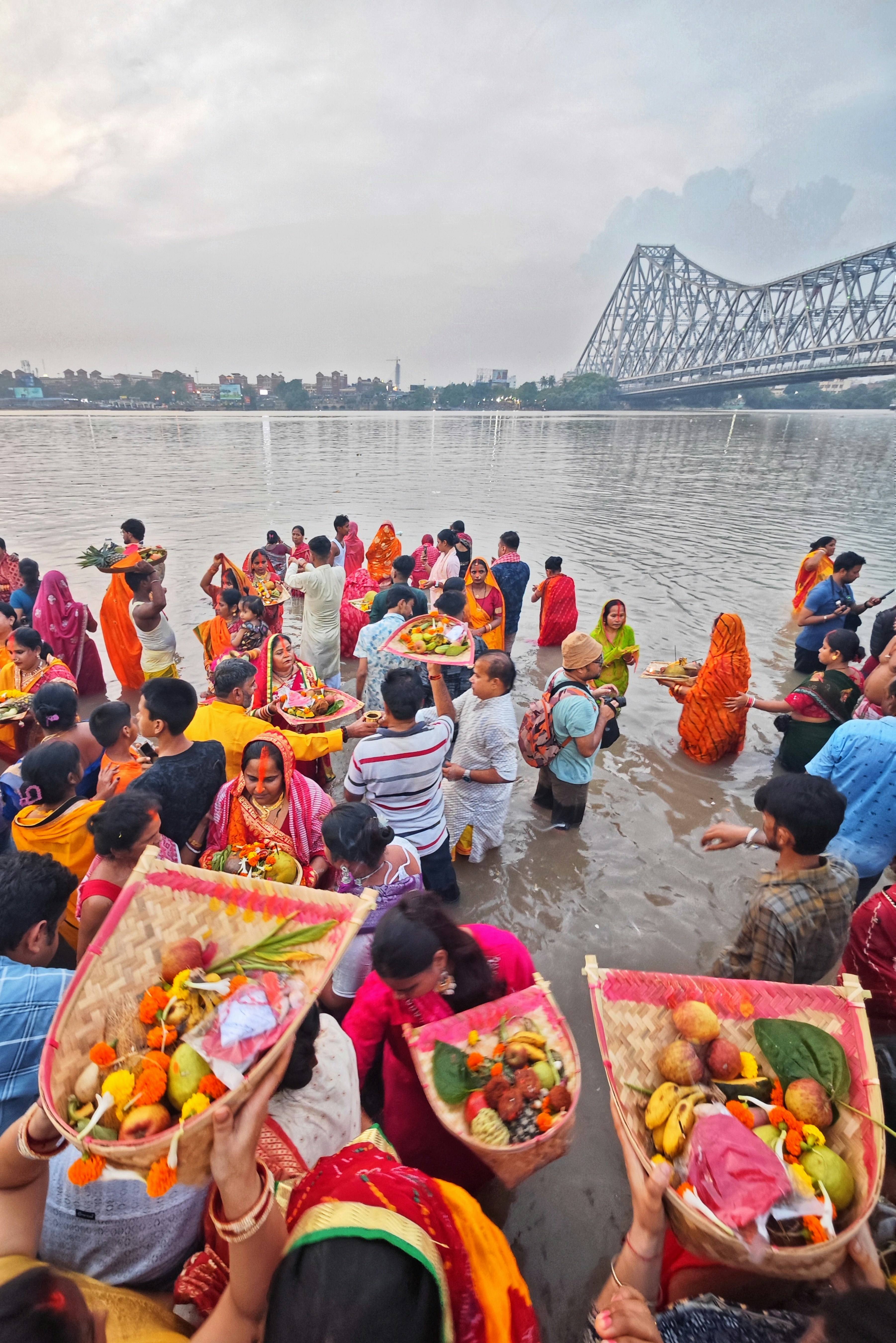 People gather by a river with offerings and a bridge.