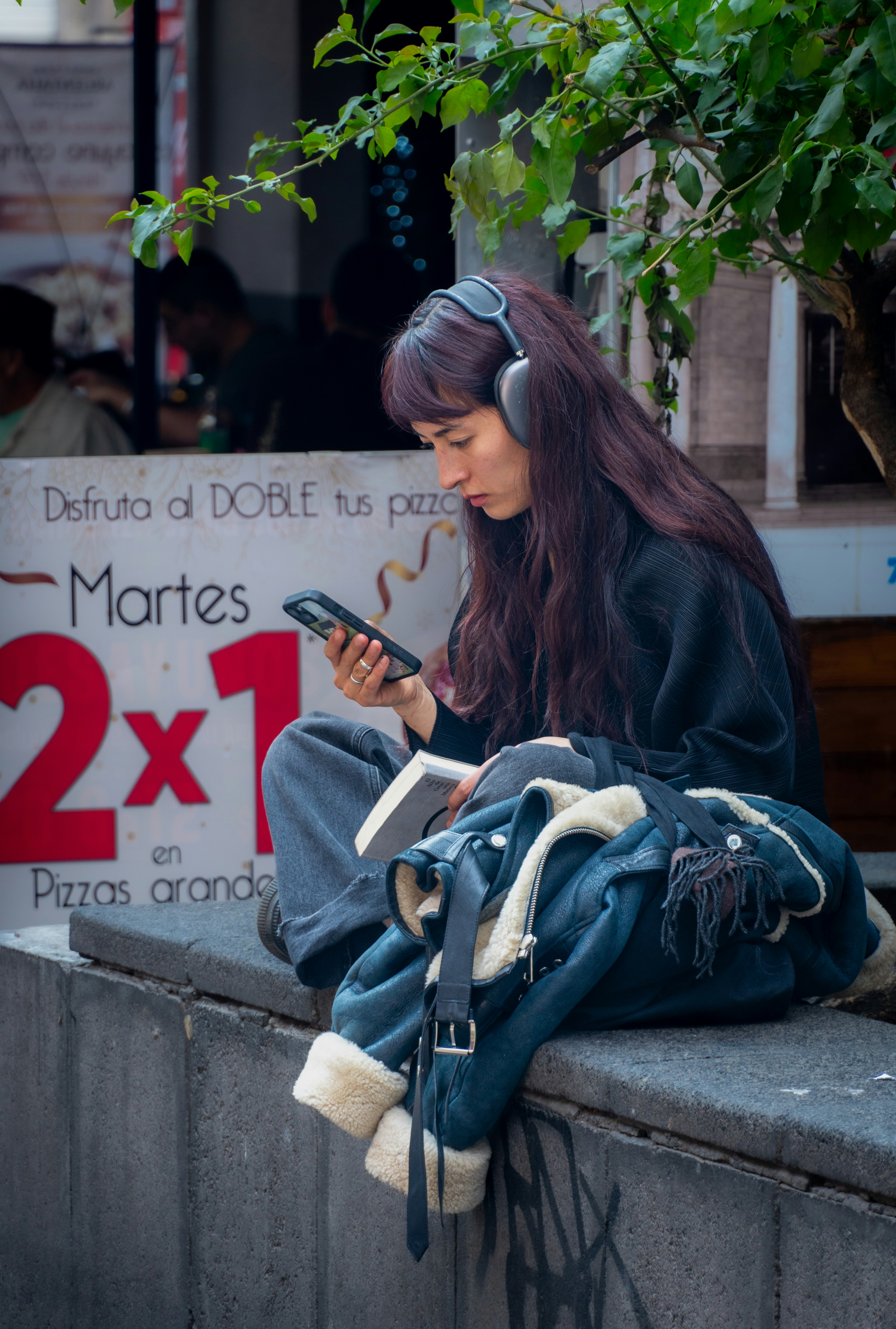 Woman with headphones reading and using phone outdoors.
