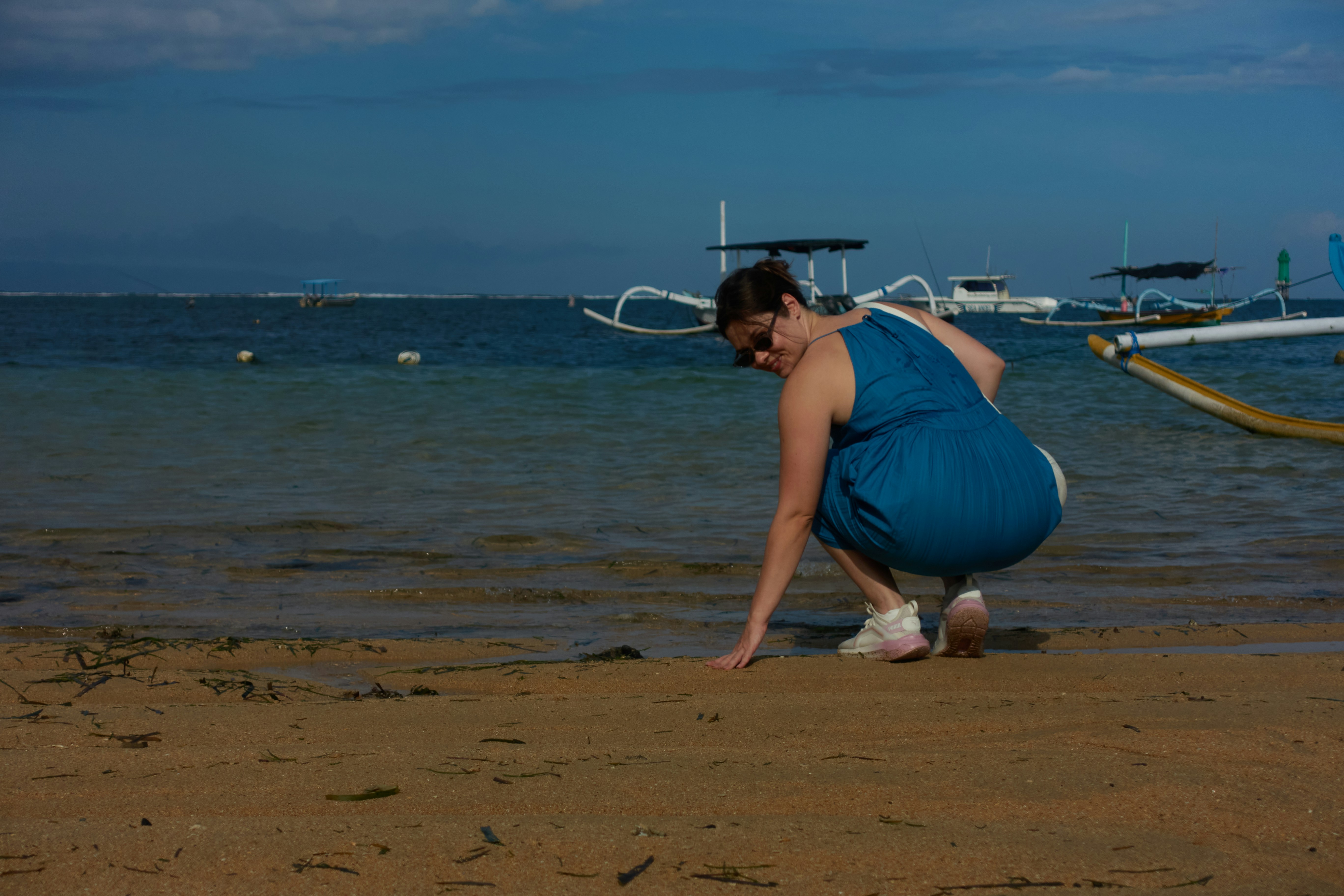 Frau im blauen Kleid, die am Strand den Sand berührt.