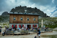 A wooden alpine mountain lodge terrace with guests dining, peaks in the background