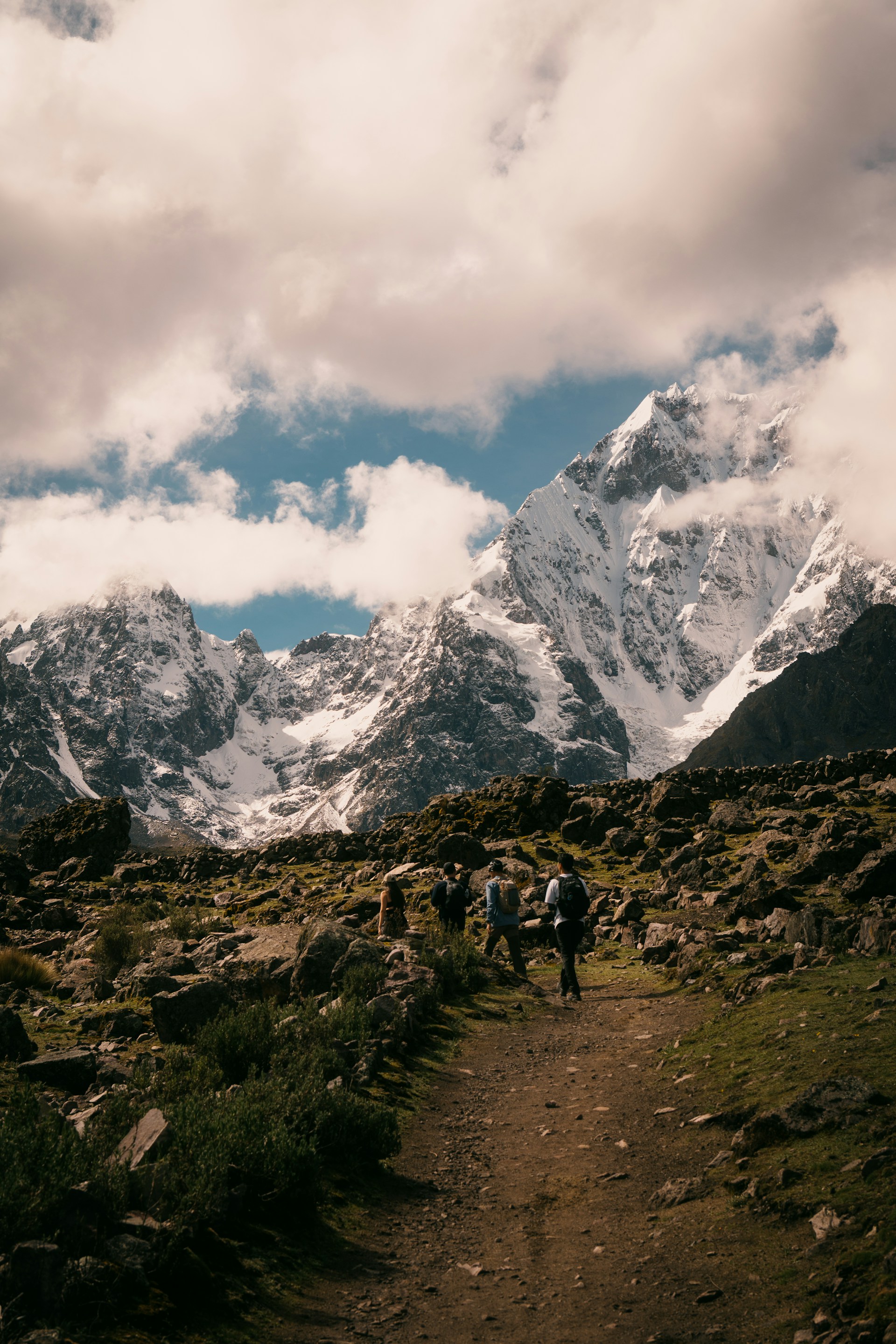 Hikers ascend a rocky mountain trail towards snow-capped peaks.