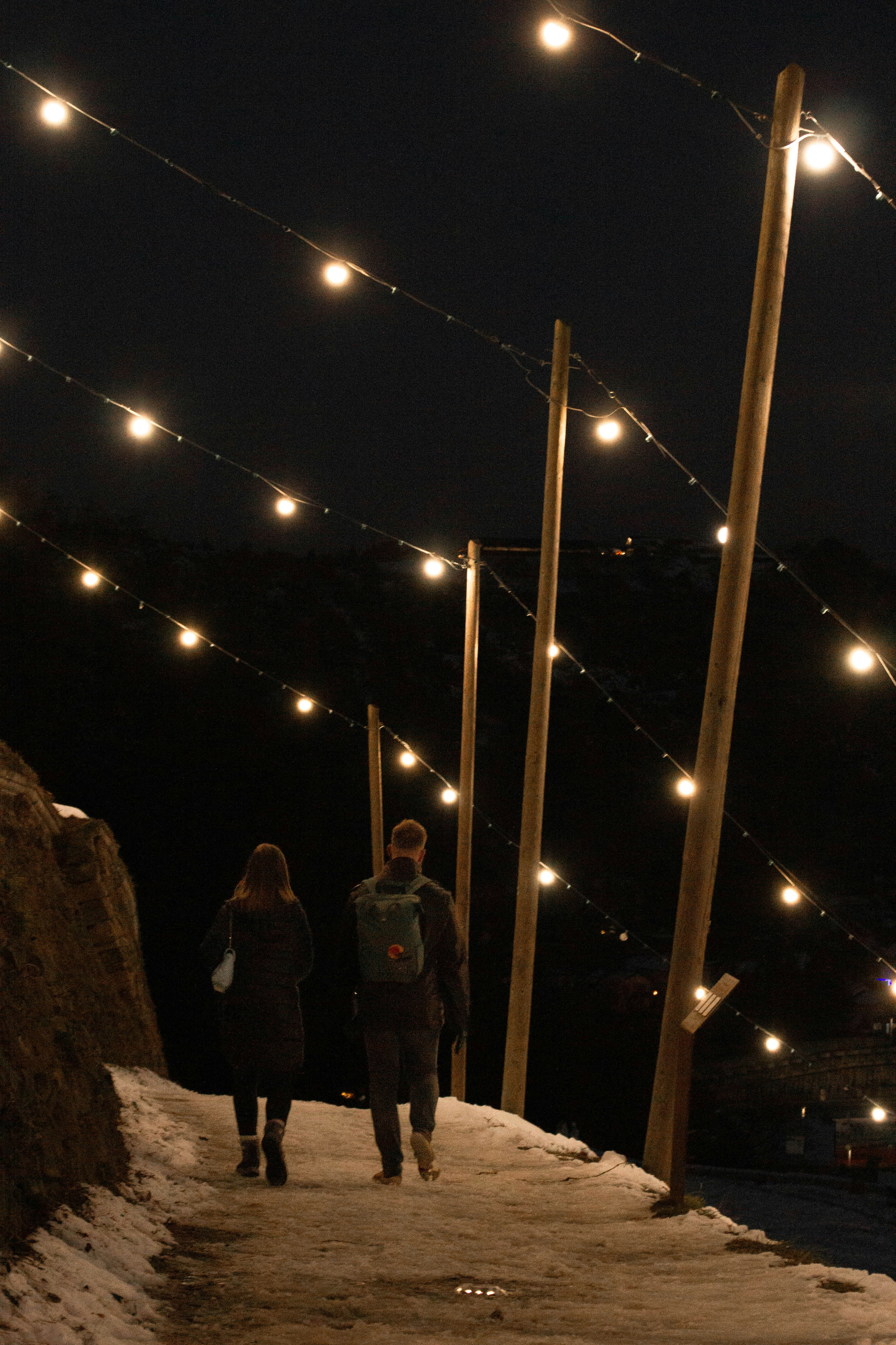 Couple walks on snowy path under string lights