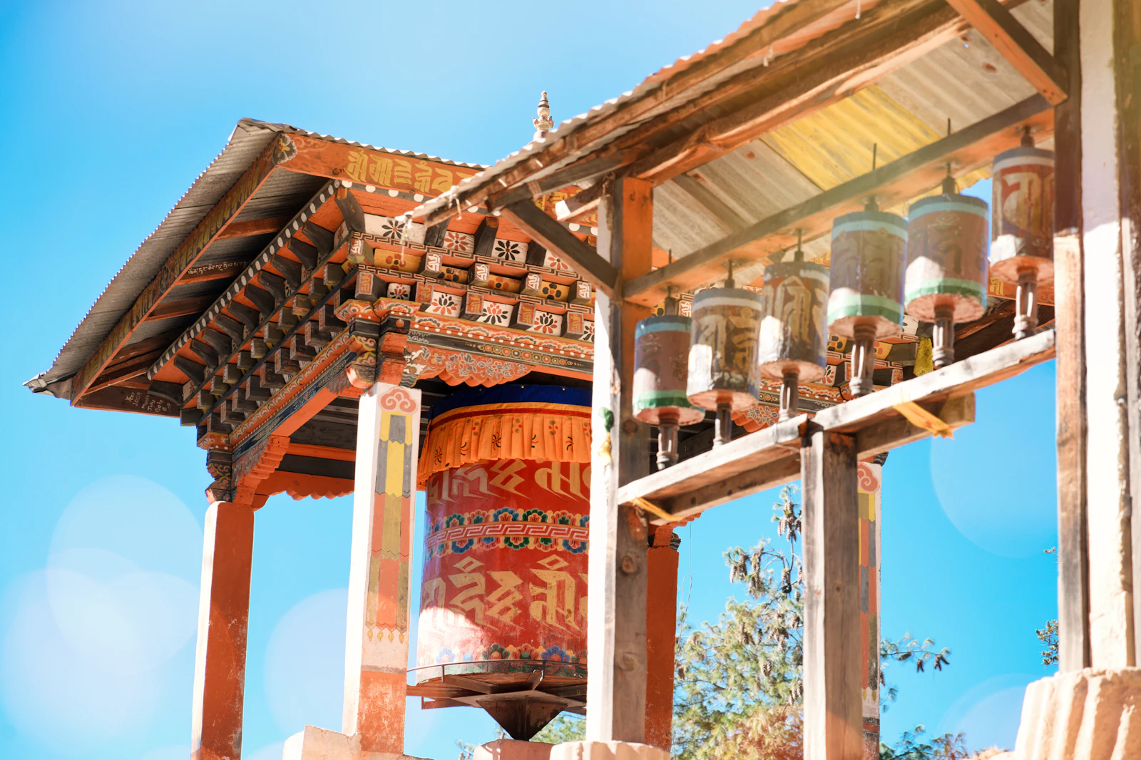 A great prayer wheel beneath a painted roof and a row of brass prayer wheels at a temple in Paro, clear blue sky
