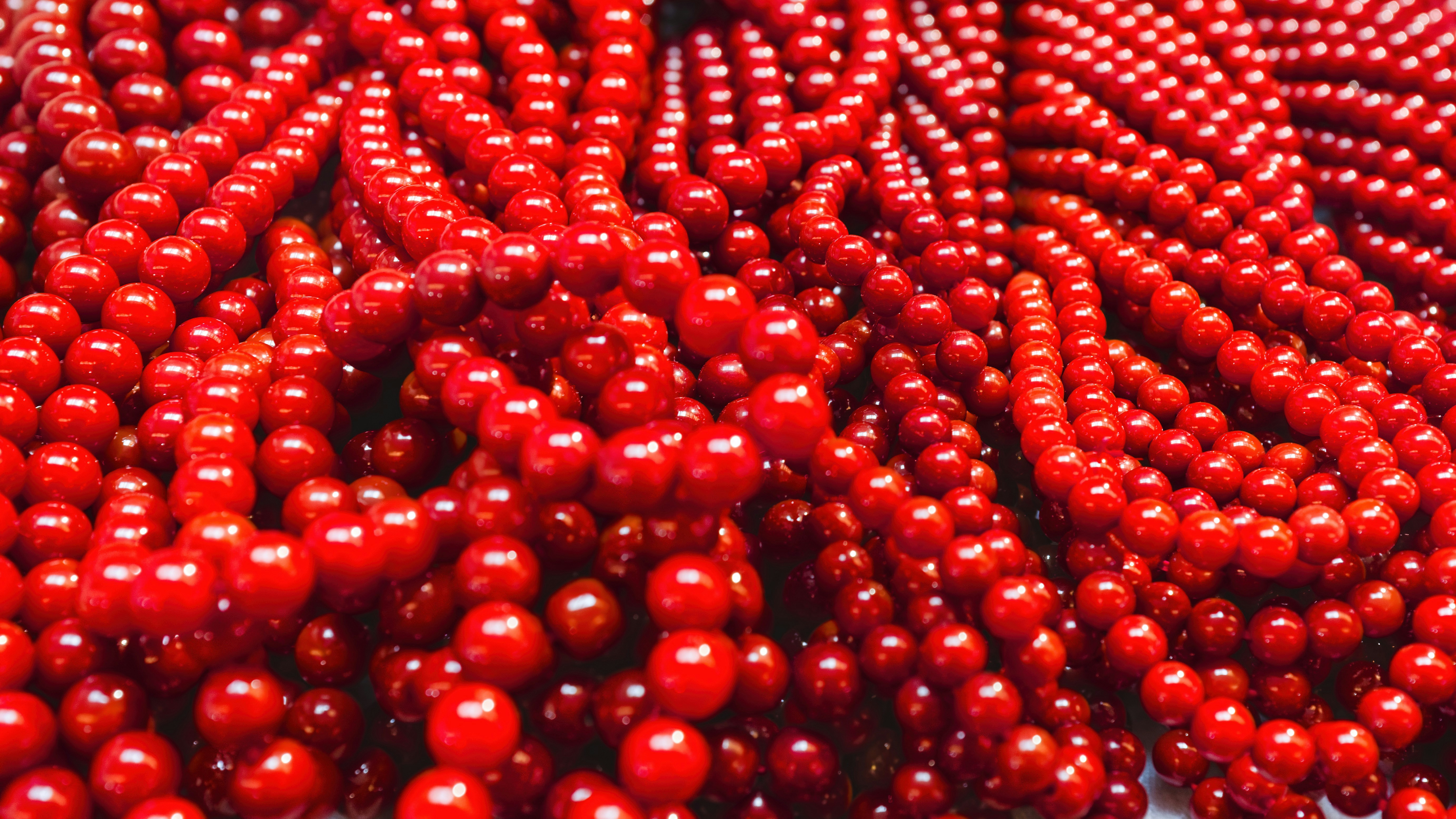 Bundles of red beads lie on a surface and create a colorful display of texture and shine in natural light during daytime - Red beads of various sizes are arranged in a random pattern on a flat surface, showing off their glossy finish. The scene is bright and vivid, capturing a close-up view of the beads.