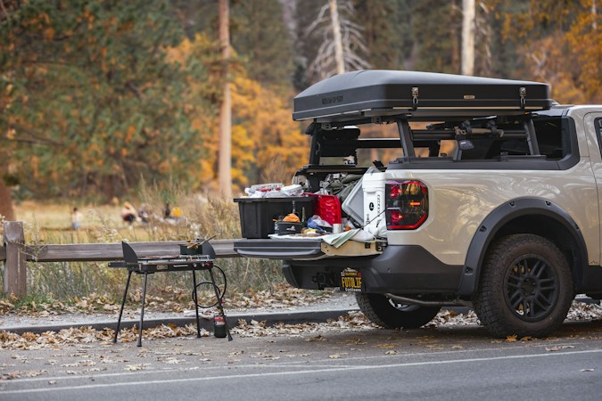 Pickup with a hard-shell rooftop tent parked beside a forest meadow in autumn, its tailgate down holding a black storage tote and cooking gear, a two-burner camp stove on legs beside it