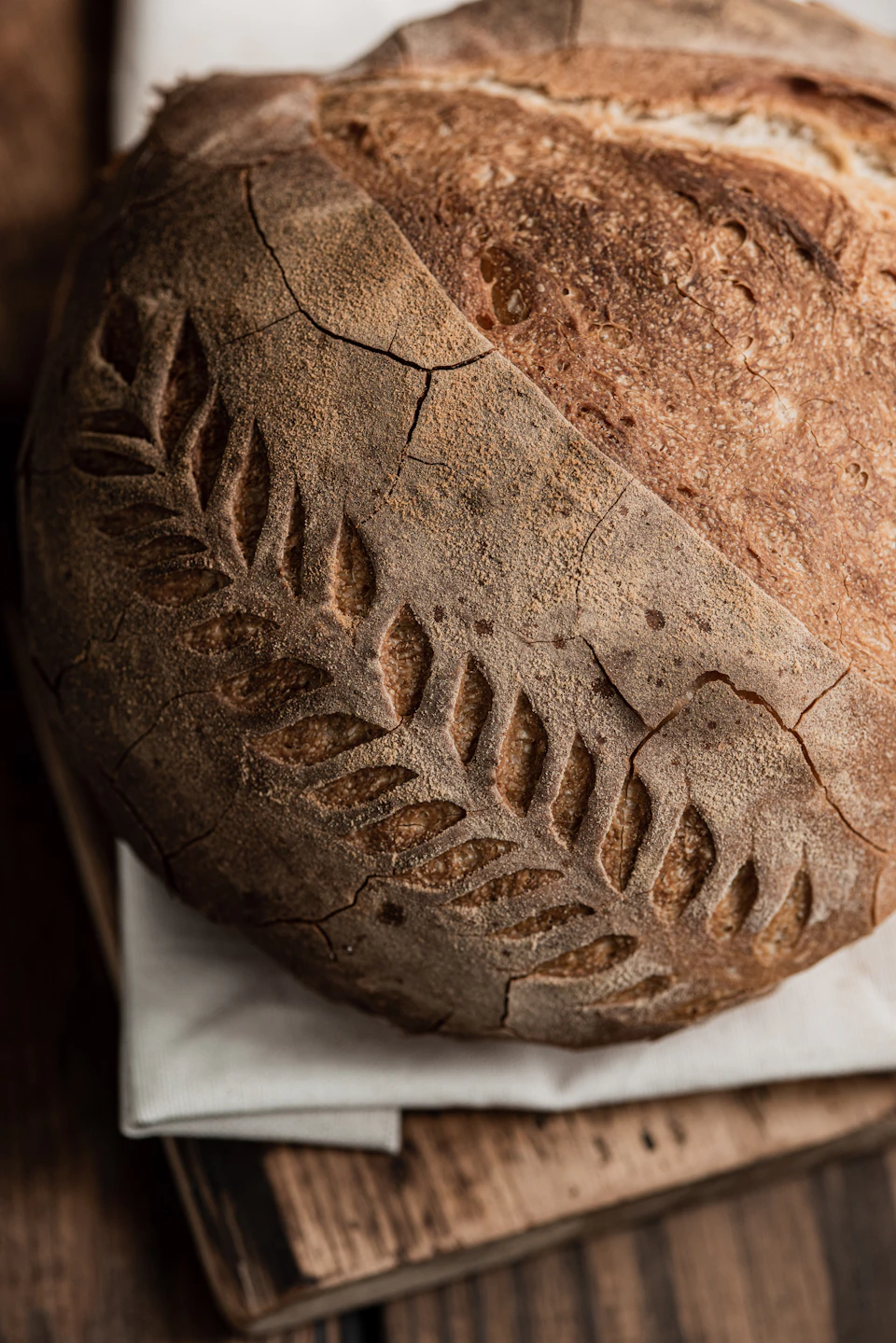 Bread with intricate leaf pattern rests on a white cloth on a wooden table