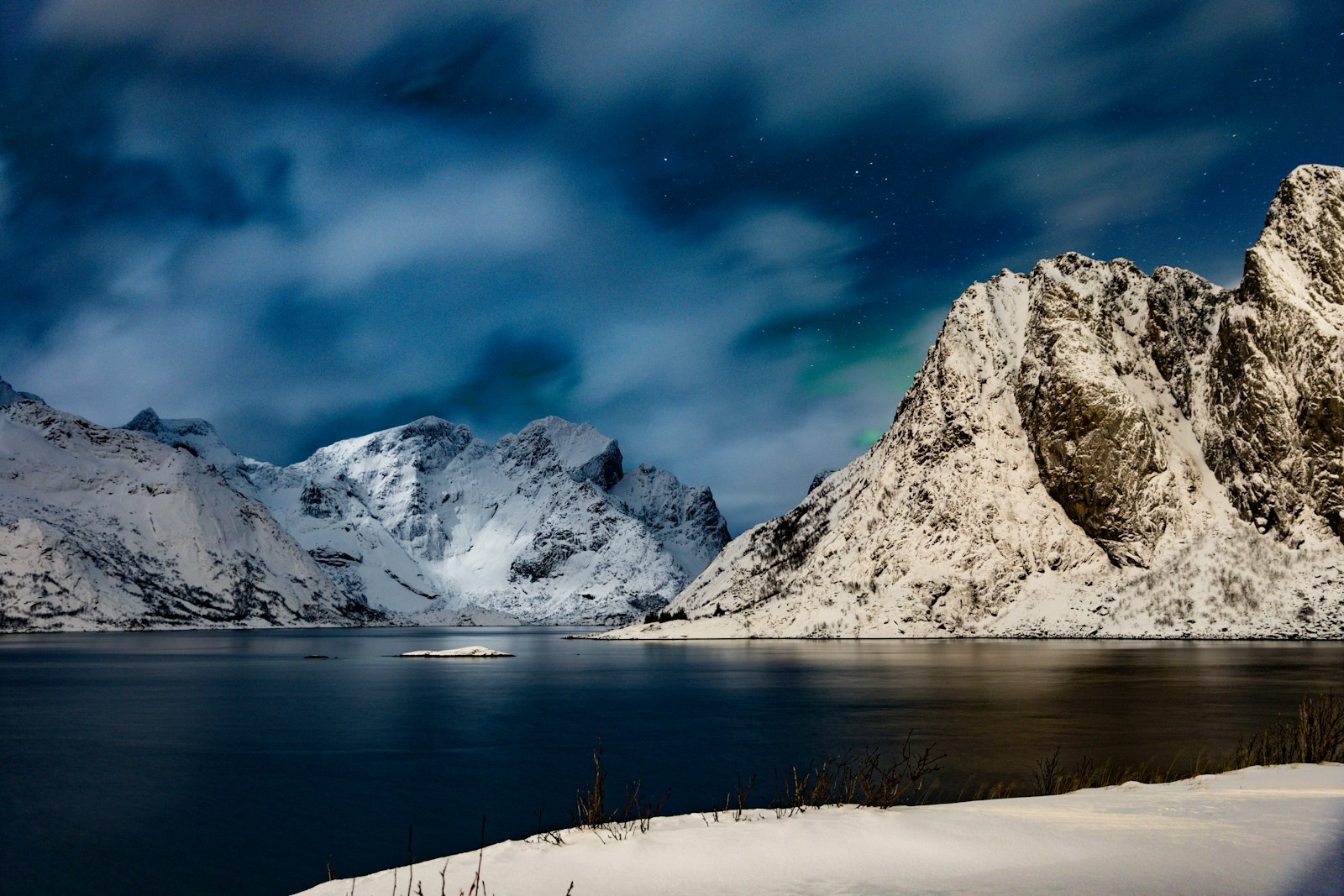 Snow-covered ridges above still water at night, with thin cloud almost entirely hiding a faint green aurora