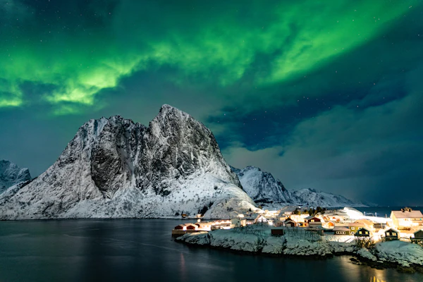 Aurora over a lit-up coastal fishing village beneath snow-covered peaks in northern Norway