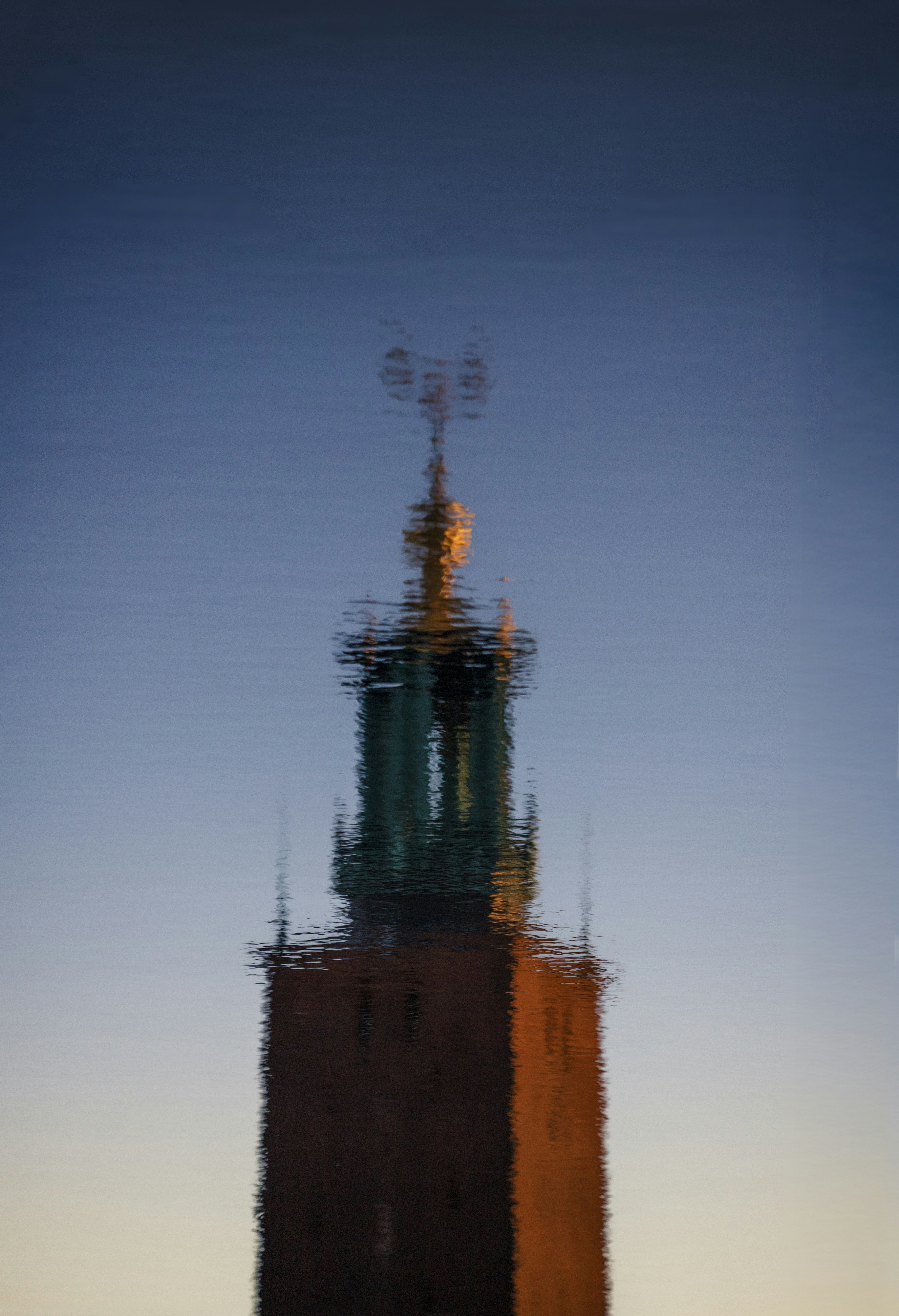 Reflection of a brick tower with a spire on water.